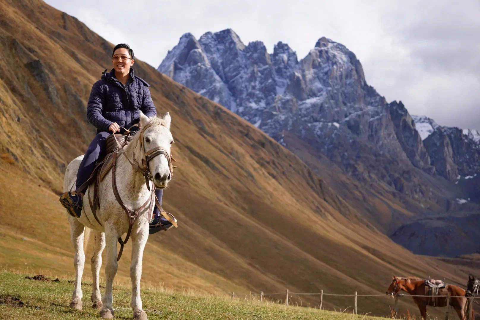 A man in a navy jacket and glasses riding a white horse in a mountainous landscape with snow-covered peaks in the background.