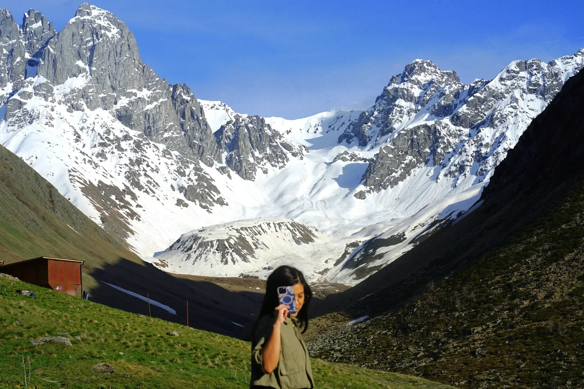 A woman with dark hair takes a photograph in a mountainous landscape with snow-capped peaks and green valleys.