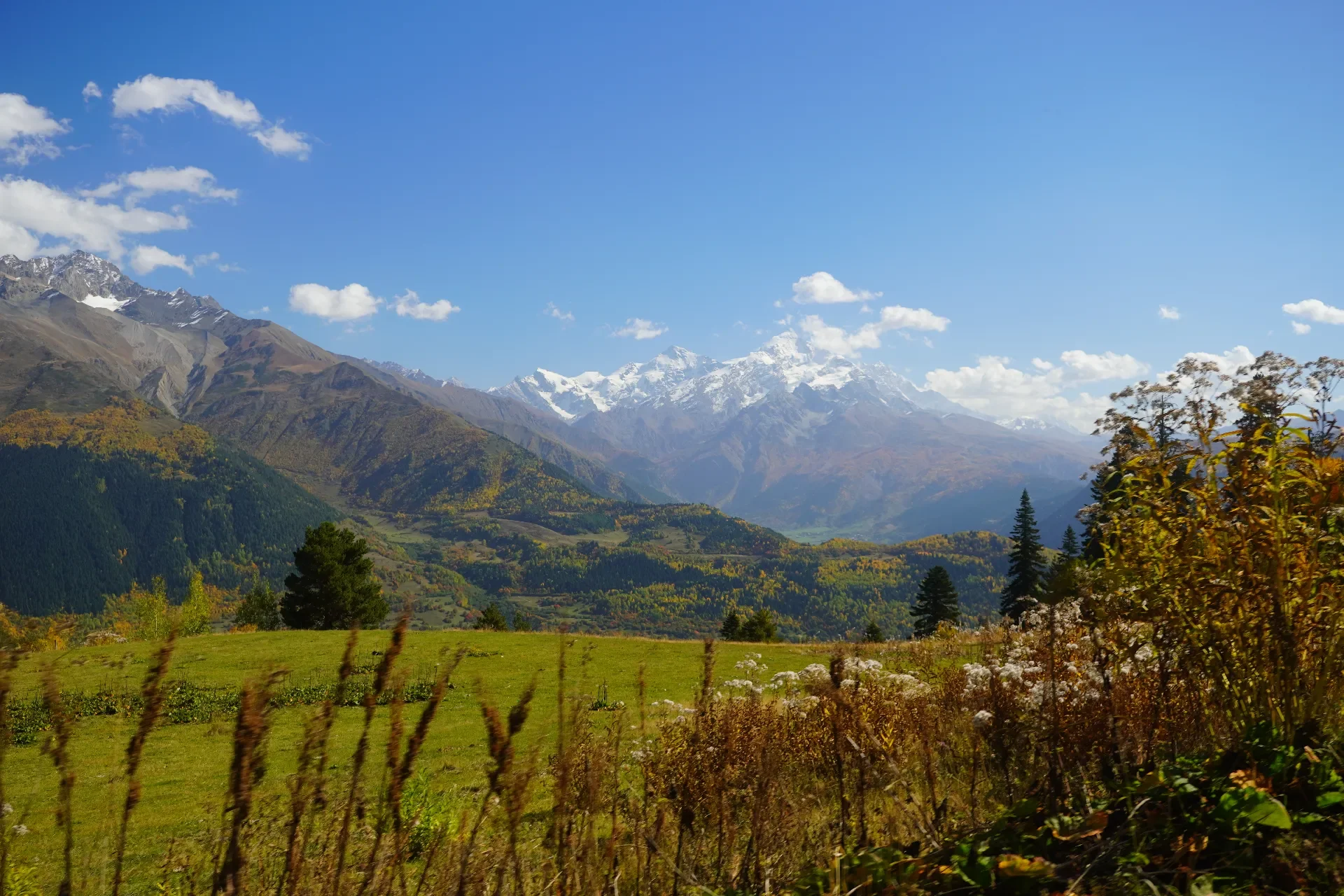Scenic view of mountains with snow caps, green and yellow lush landscape, and clear blue sky with clouds.