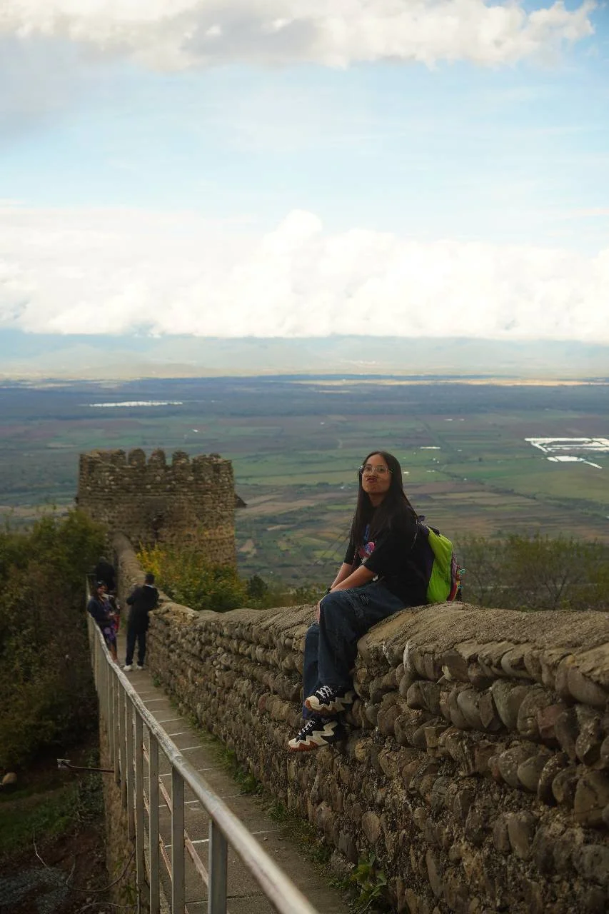 A young woman sitting on an ancient stone wall atop a hill with a scenic view of the landscape behind her, including fields, water bodies, and a cloudy sky.
