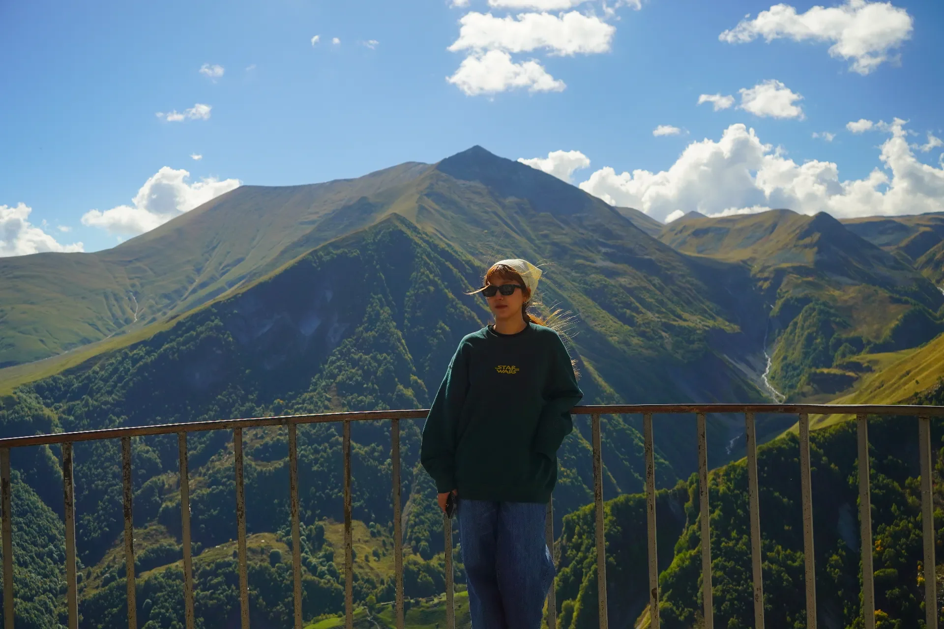 A woman wearing sunglasses, a bandana, a green Star Wars sweatshirt, and jeans standing behind a metal railing, with green mountains and a blue sky with clouds in the background.