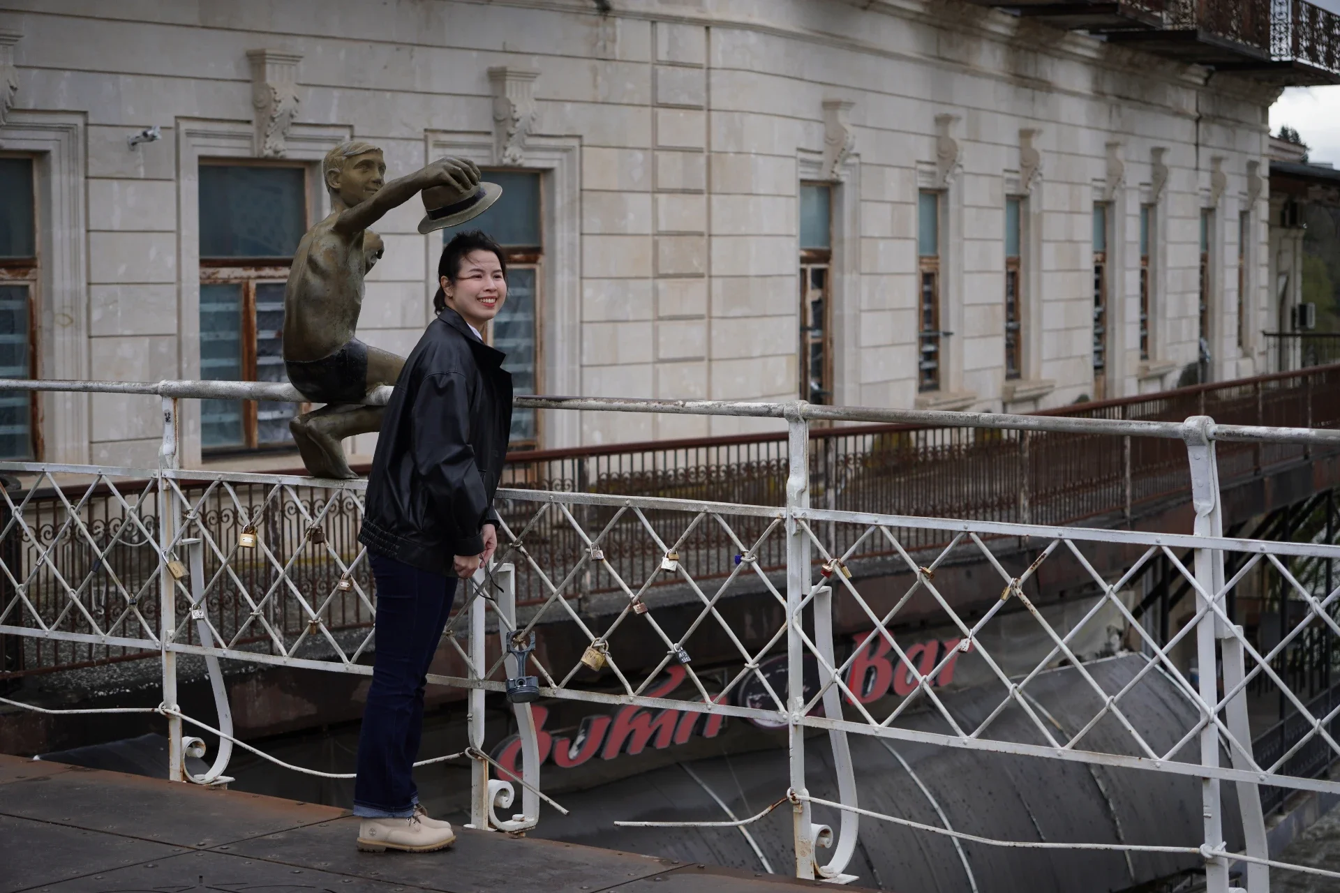 A young woman with dark hair smiling and wearing a black jacket, jeans, and beige boots standing near a white metal fence with padlocks, in front of a historic building with cream-colored stone facade, multiple windows, and a decorative balcony, unde