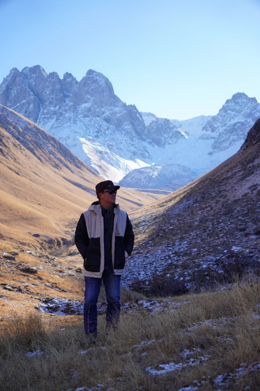 A man stands in a mountainous landscape with snow-capped peaks in the background. He wears sunglasses, a black cap, and a layered jacket, looking off into the distance.