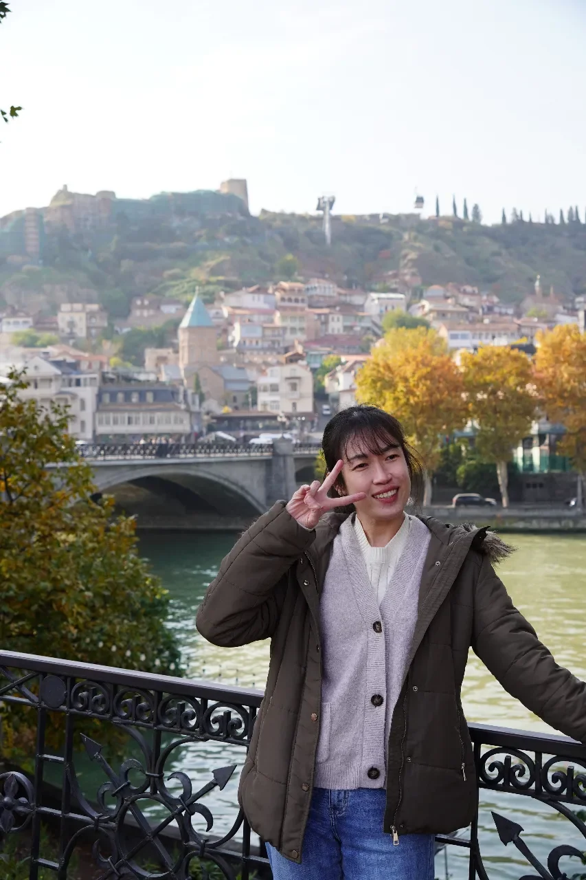 A young woman standing by a river in a city park, smiling and making a peace sign with her hand, with a historic bridge, colorful trees, and a town on a hill in the background.