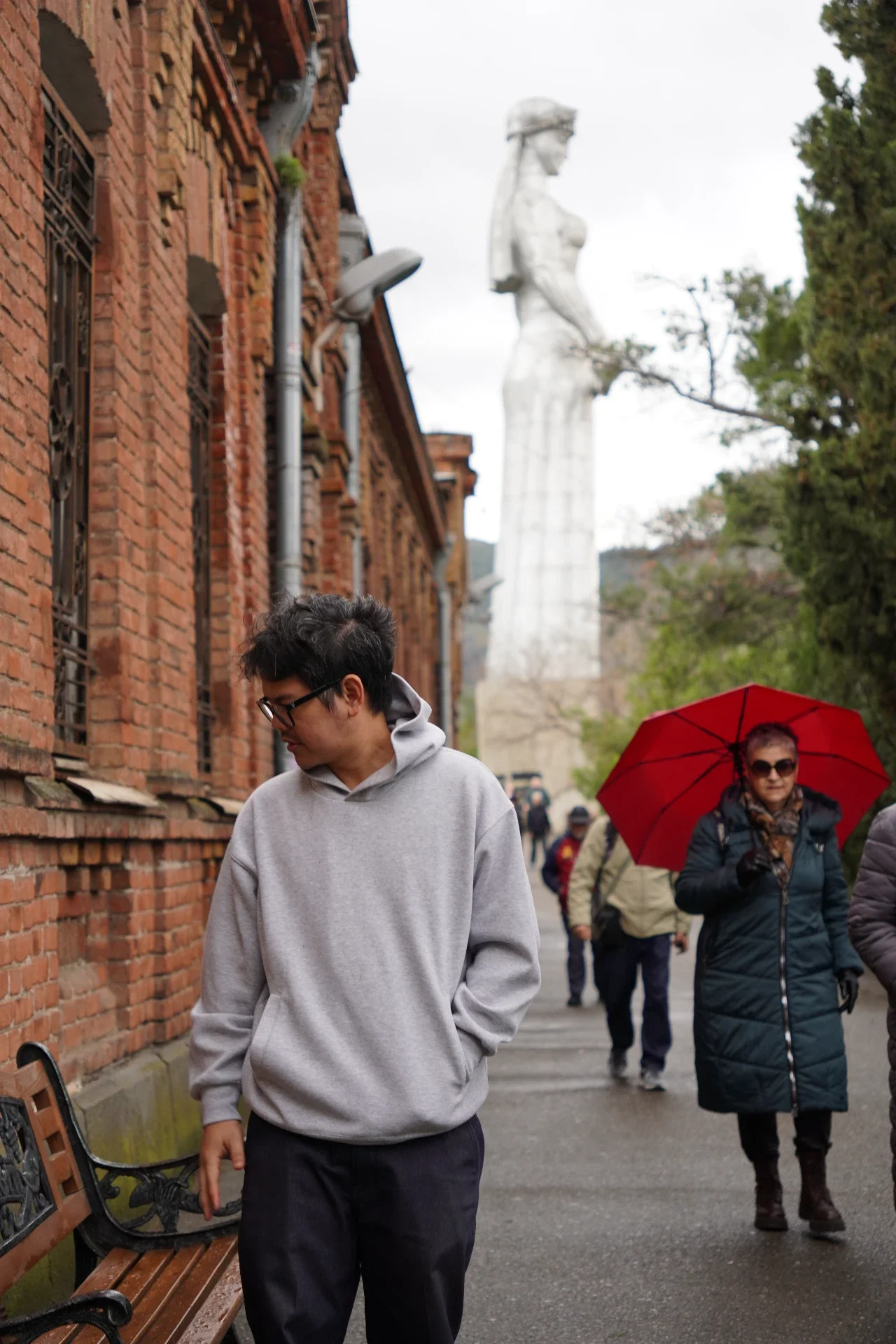 People walking along a sidewalk next to a brick building, with a large statue of a woman in the background, some trees, and a woman holding a red umbrella.