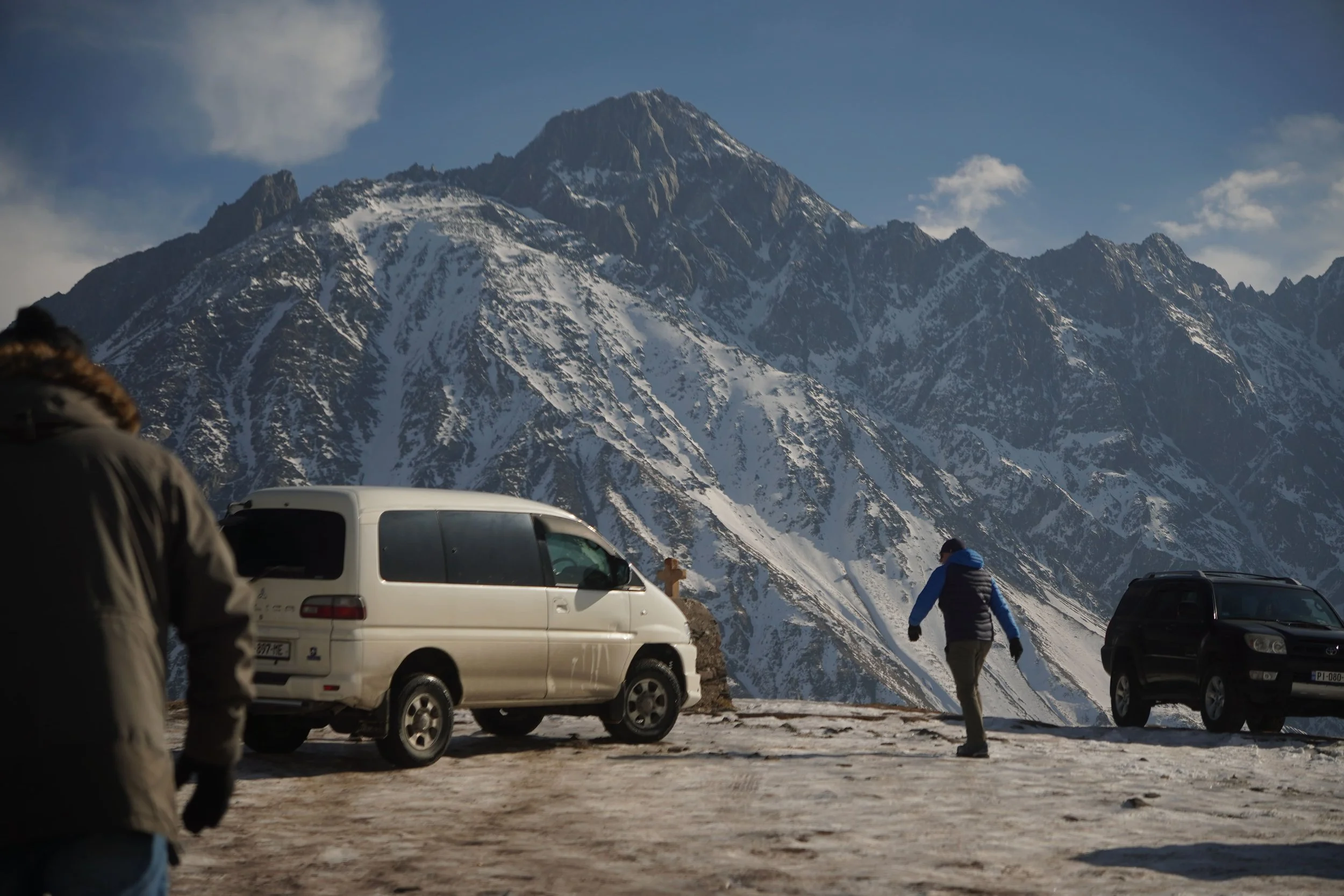 Snow-covered mountain with two cars and three people in winter clothing standing on a mountain road.