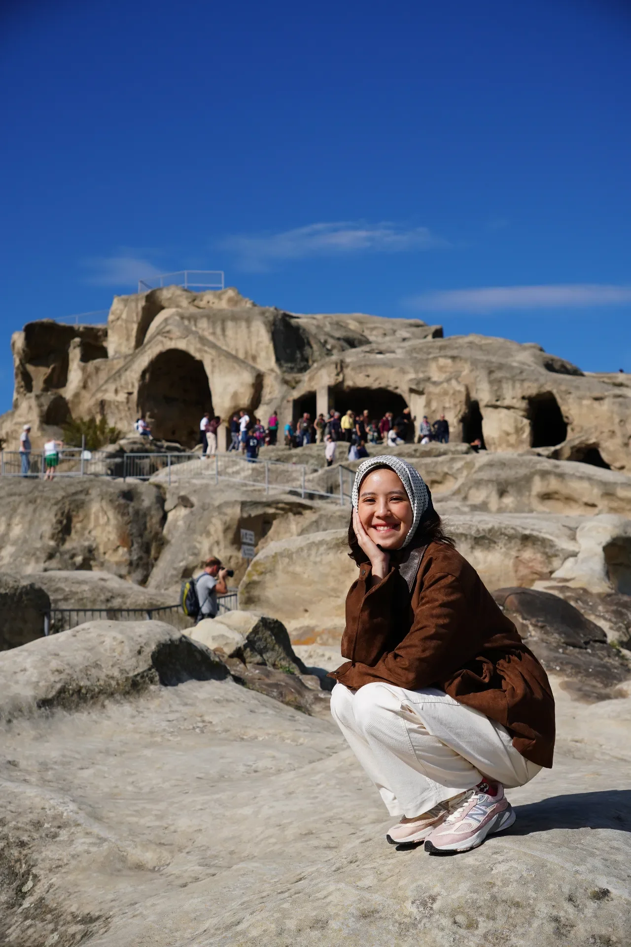 A young woman squatting on rocky terrain, smiling, dressed in a brown jacket, white pants, and pink sneakers, with a historic site and a crowd of people in the background under a bright blue sky.
