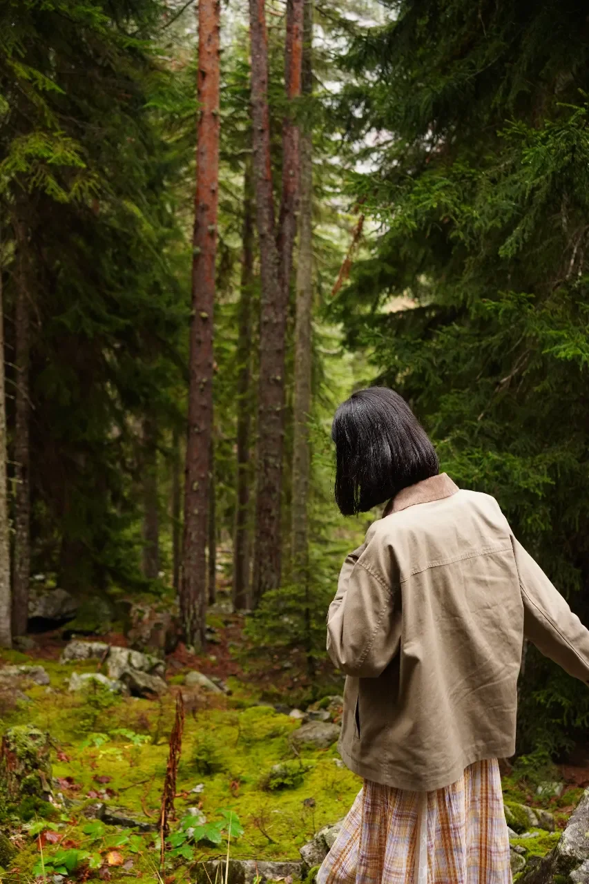 Woman with black hair in a beige jacket and plaid skirt walking in a dense green forest.