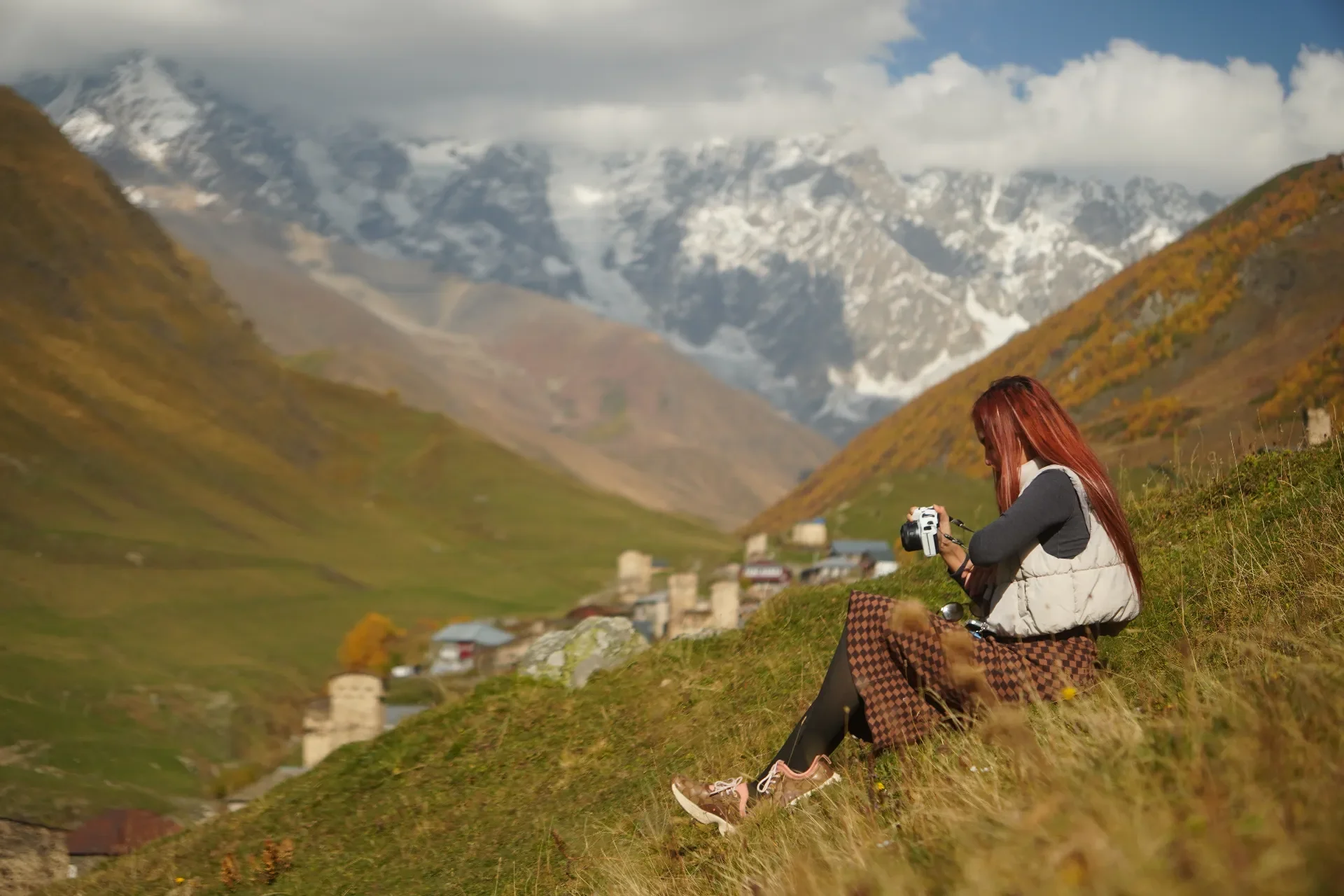 A woman with red hair sitting on a grassy hill, holding a camera, with mountains and a village in the background.