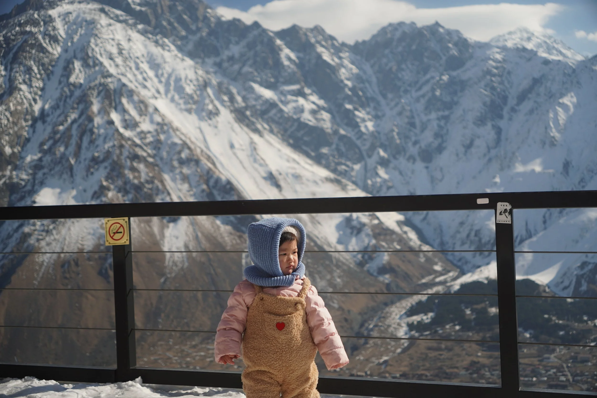 Young child in pink winter coat and teddy bear costume standing on snow-covered ground at a mountain viewing platform, with snow-capped mountains in the background.