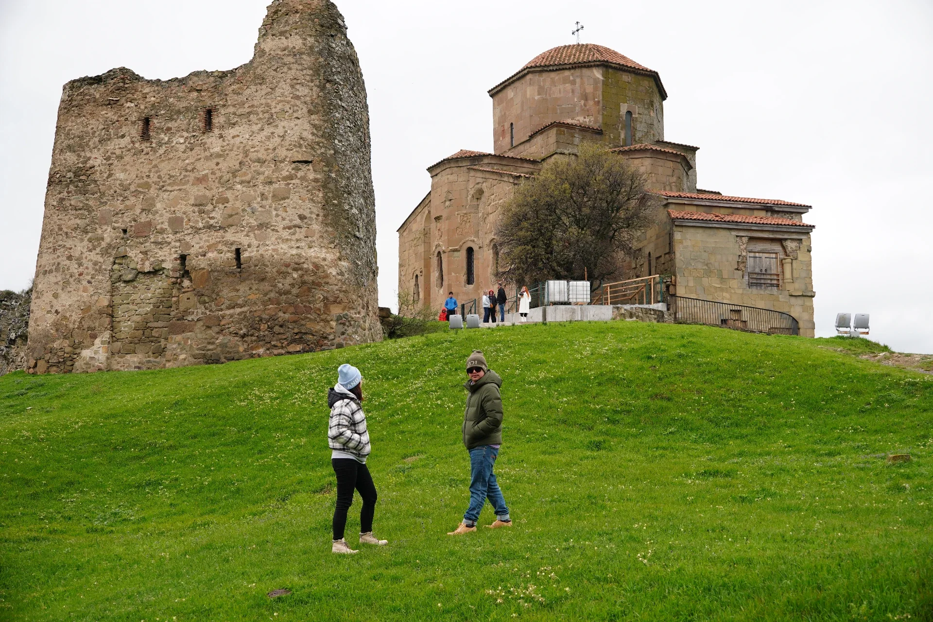 People standing on a grassy hill near an old stone church with a small tower and a round dome. The sky is overcast.