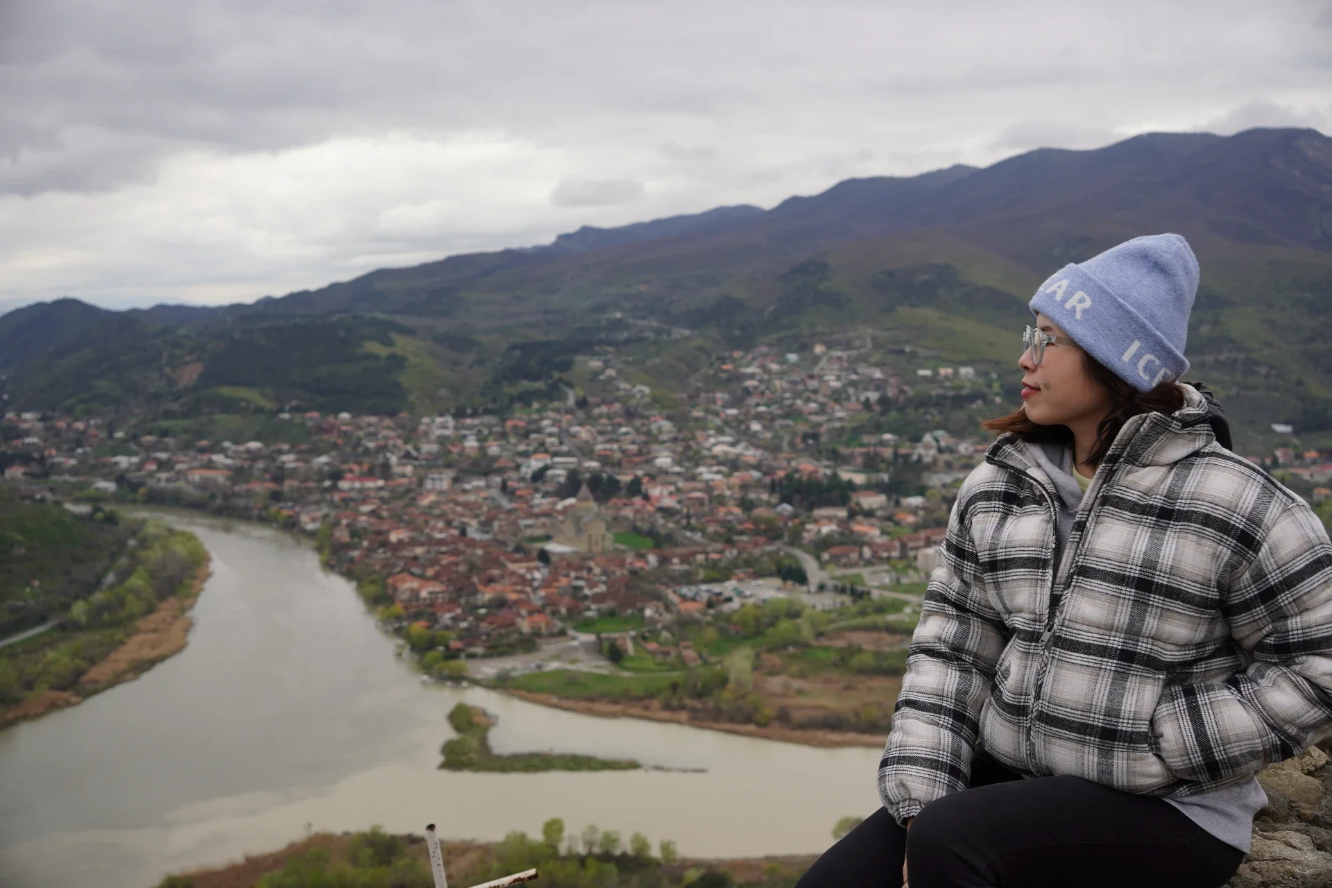 A woman wearing a beige and black plaid jacket and a blue knit hat sits on a mountain overlooking a town with a river running through it, surrounded by green hills and mountains under a cloudy sky.