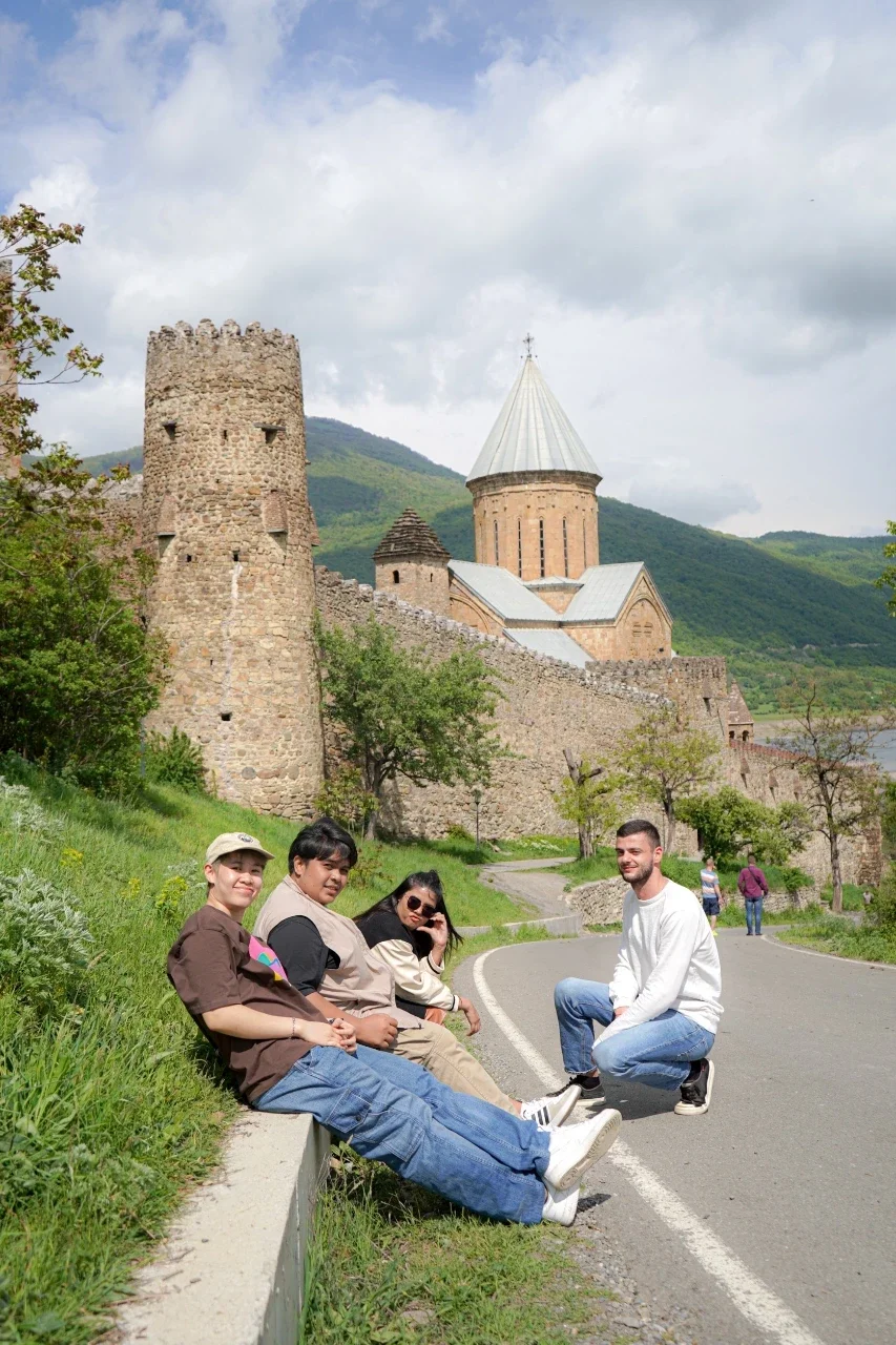 A group of four young people sitting and standing on a curved road with an ancient stone castle and green hills in the background.