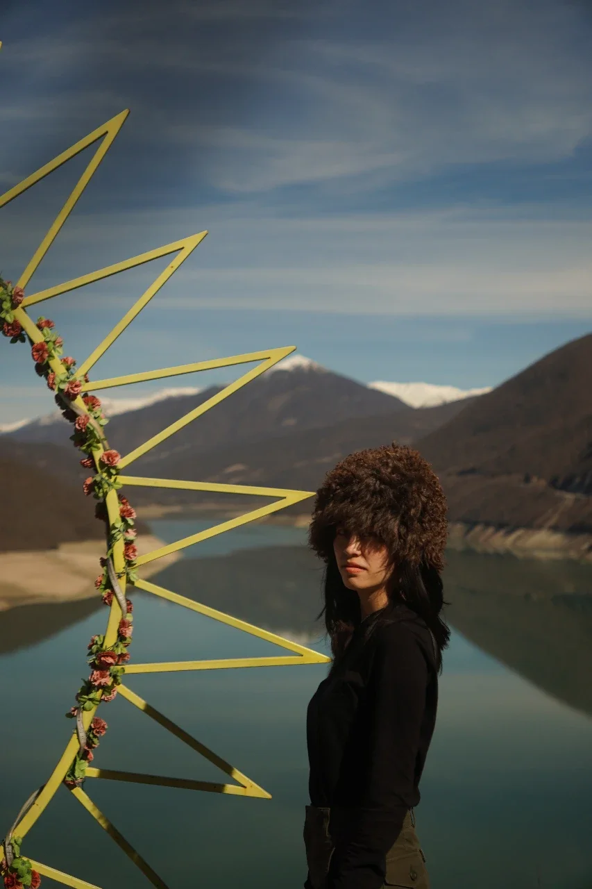 A woman wearing a large furry hat stands outdoors near a yellow geometric sculpture decorated with flowers, with a mountains and lake landscape in the background.