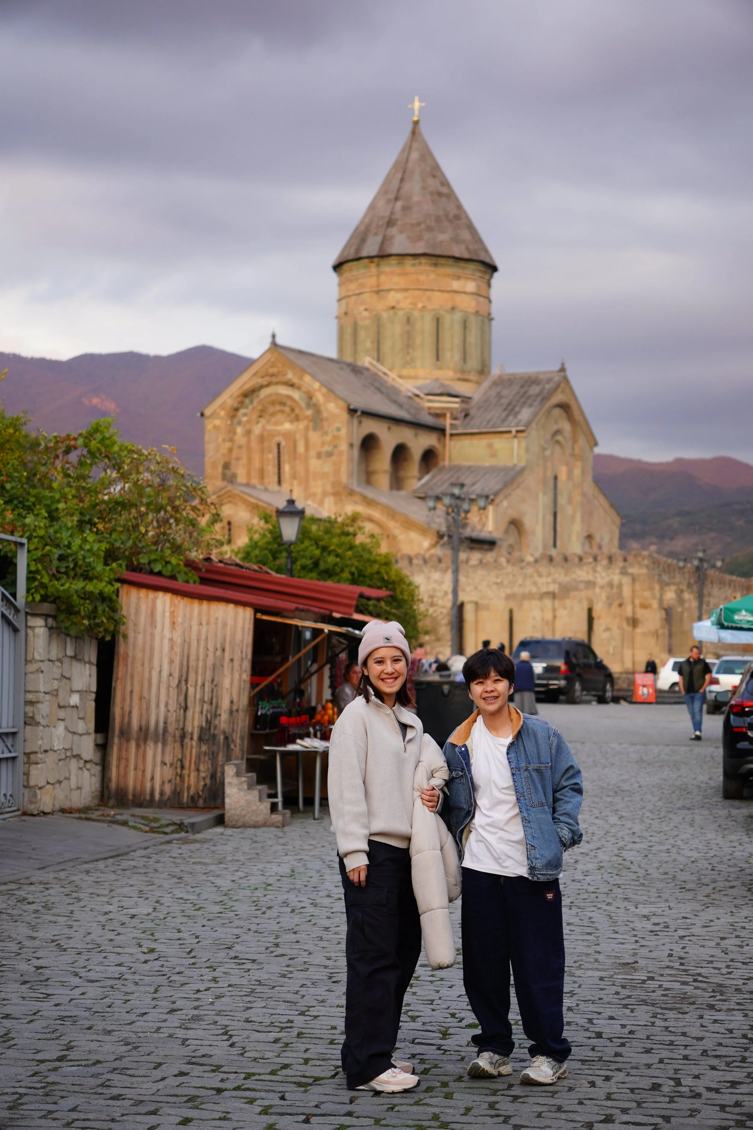 A woman and a man standing on a cobblestone street in front of a historic brick church with a stone dome and cross on top, with mountains in the background.