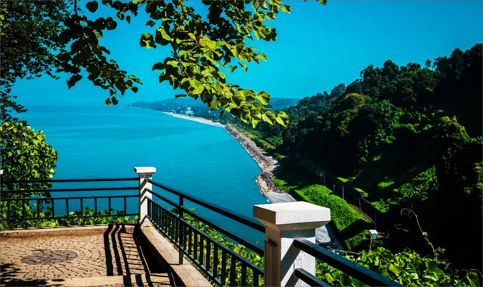 View overlooking a blue lake and green hills from a lookout with a black metal railing and stone posts, surrounded by leafy trees.