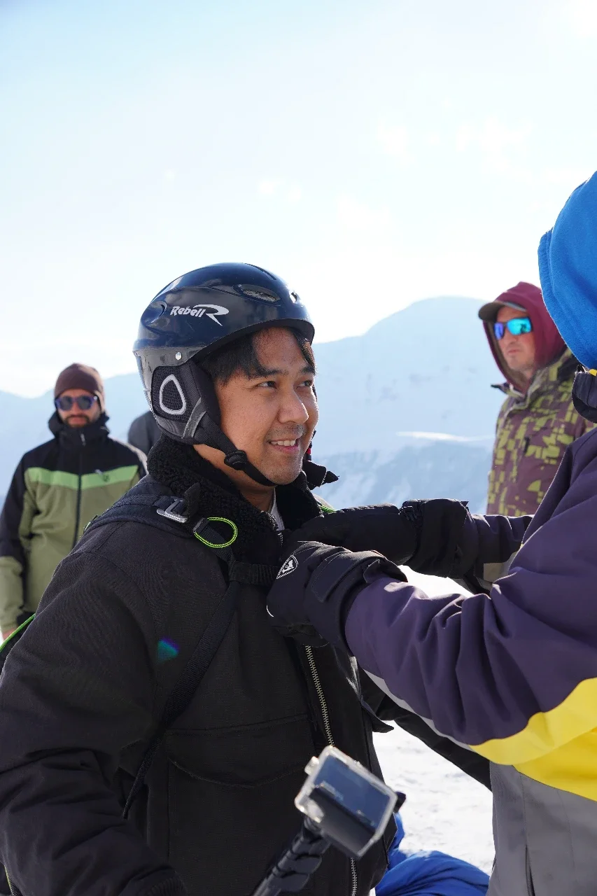A man in a black jacket and ski helmet smiling as another person adjusts something on his jacket, with snow-covered mountains and other people in winter gear in the background.