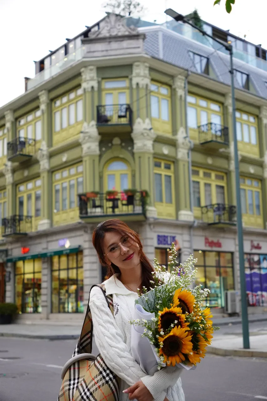 A young woman holding a bouquet of sunflowers and white flowers, smiling, standing on a city street in front of a colorful old building with ornate architecture and multiple balconies.