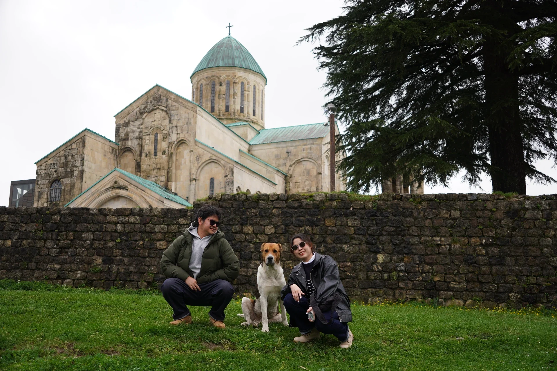 Three people and a dog sitting on a grassy area in front of a brick wall with a historic church and large tree in the background.