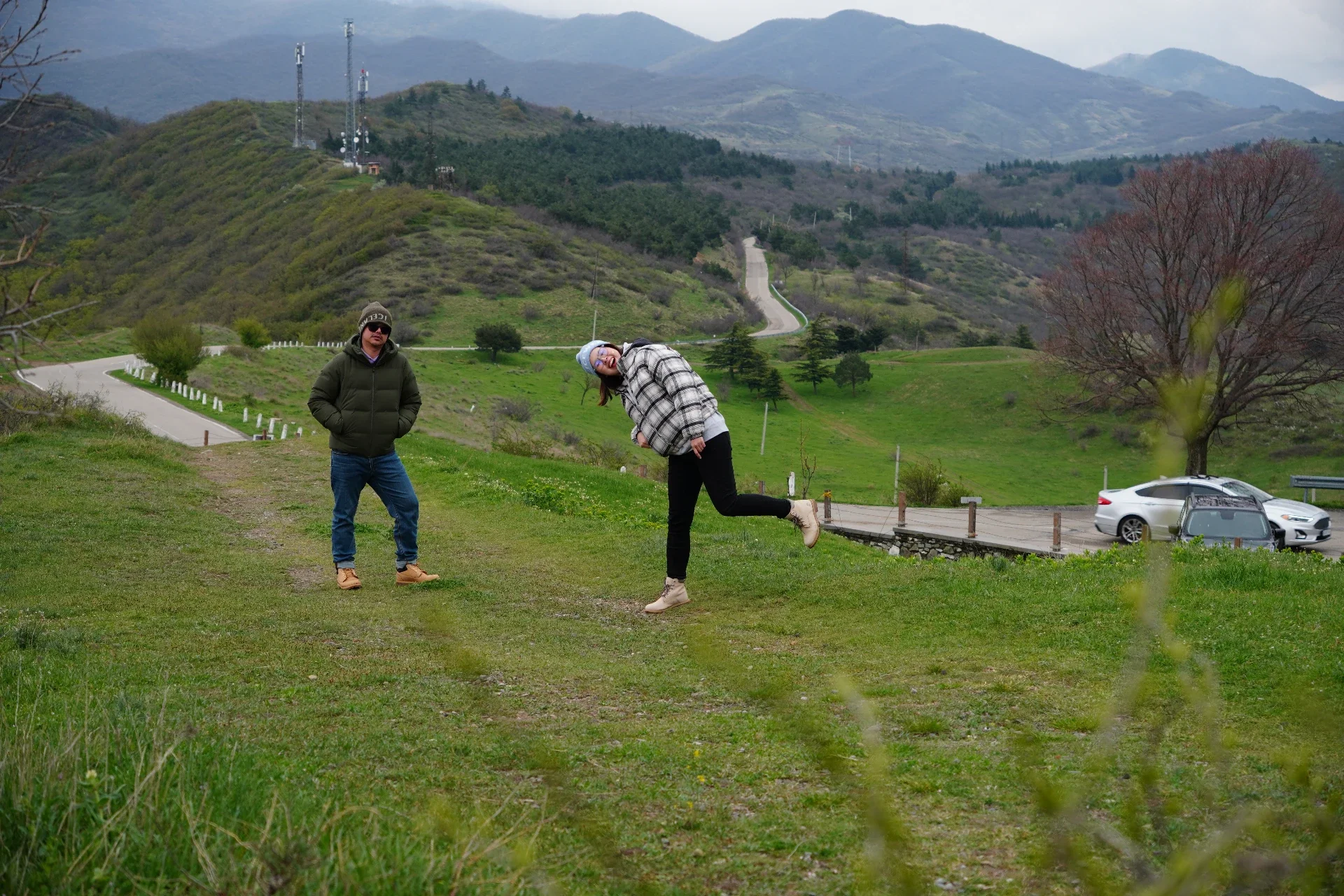 Two people standing on a grassy area in a hilly landscape, with mountains and trees in the background. One person is wearing a green jacket, sunglasses, and a beanie, standing with hands in pockets. The other person is wearing a plaid jacket, a gray 