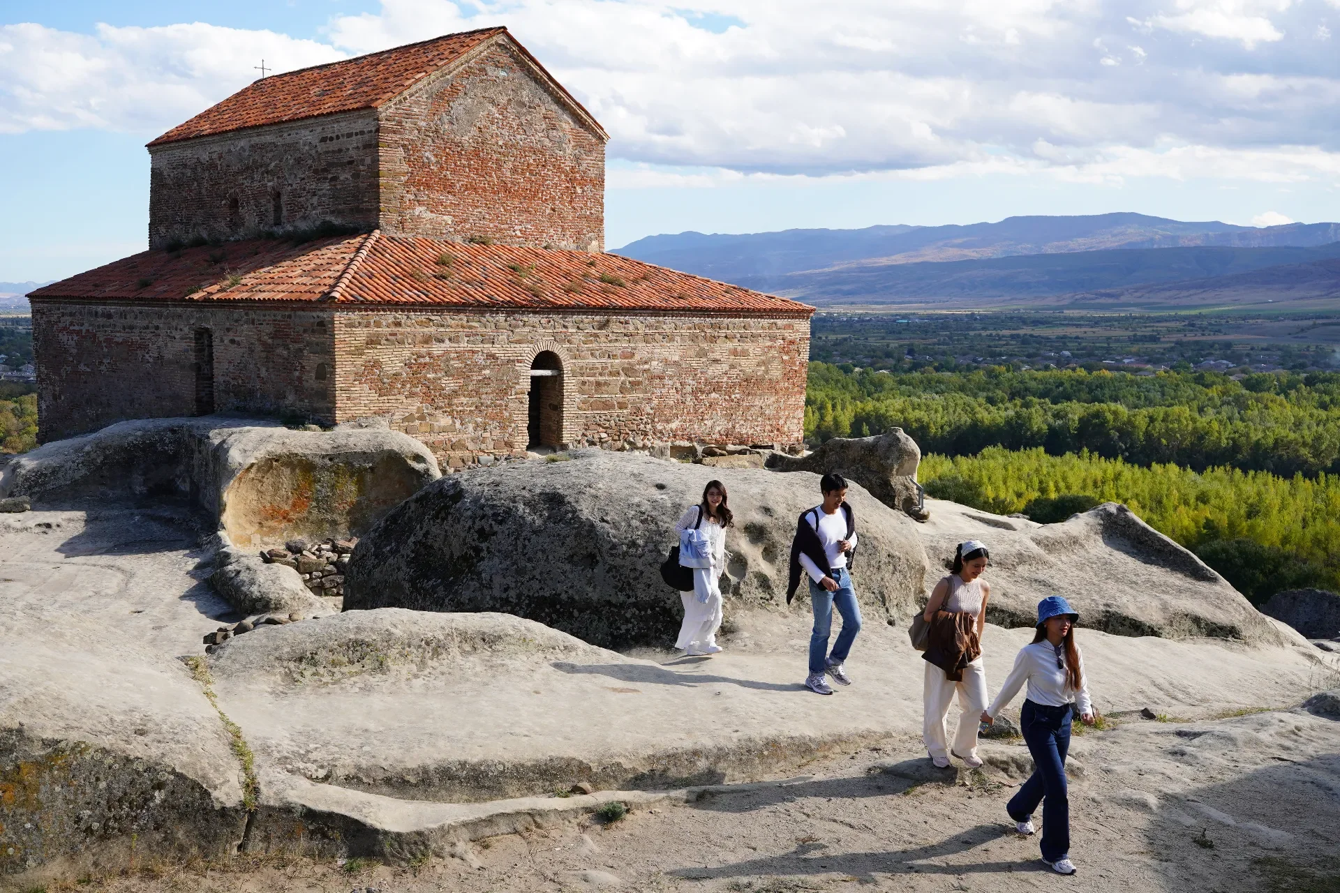 Group of five people walking on rocky terrain in front of an ancient stone building with a brick roof, with a scenic landscape of green trees and mountains in the background.