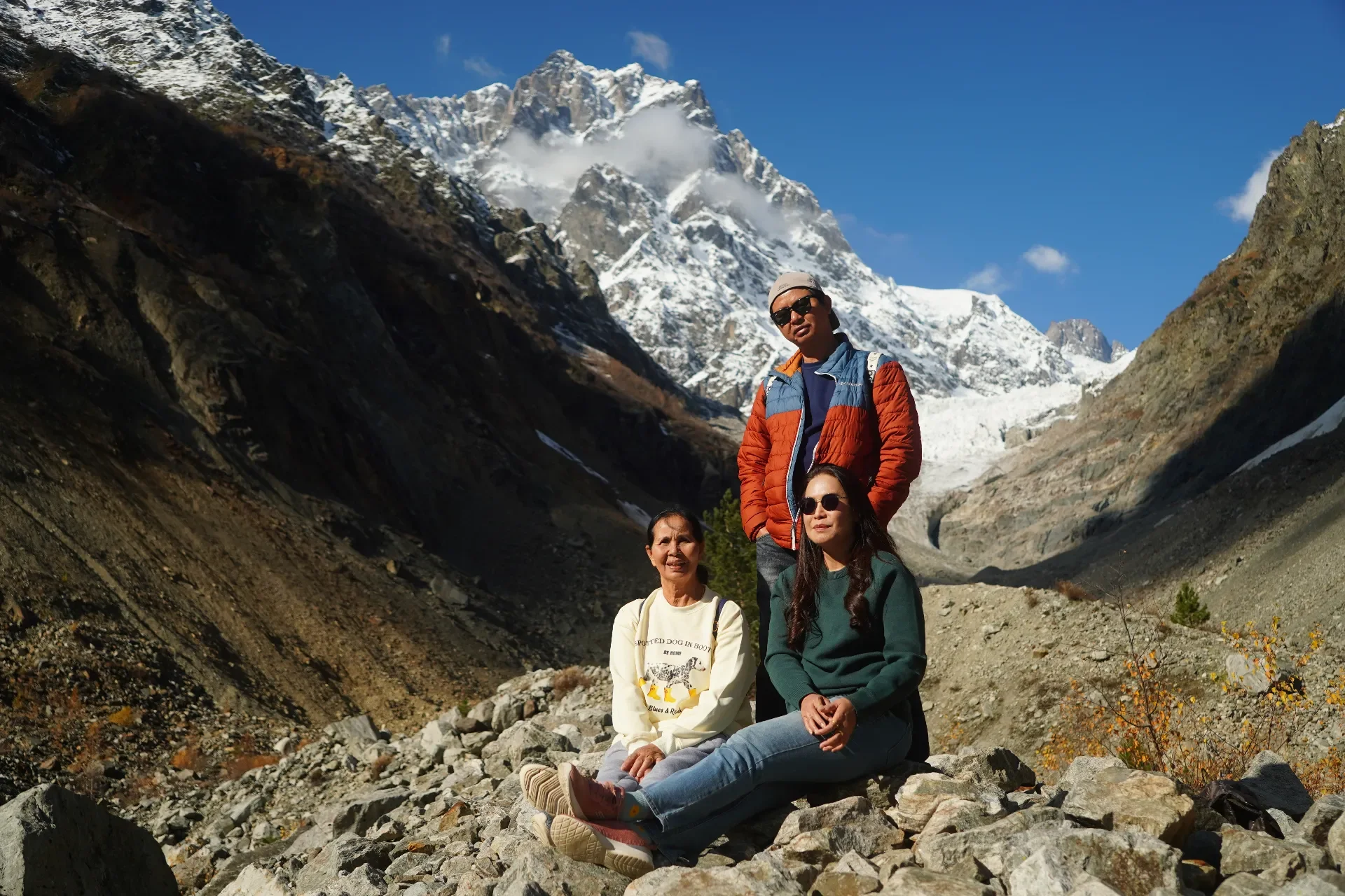 Three people hiking in a rocky mountain valley with snow-capped peaks in the background. One person is standing, wearing a colorful jacket and sunglasses; two others are sitting on rocks, wearing casual outdoor clothing.