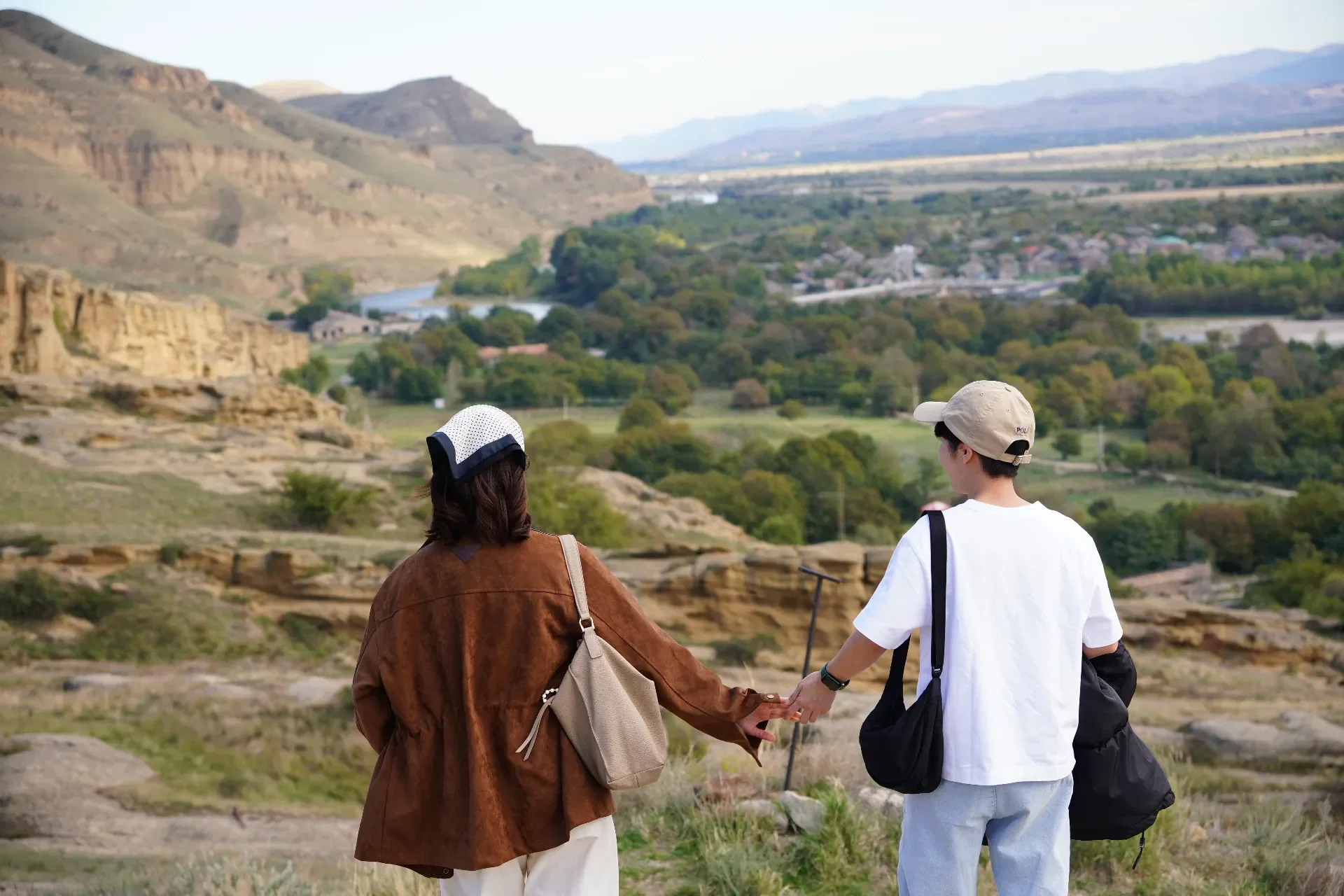 A woman and a man holding hands while walking in a scenic outdoor landscape with green hills and a river in the background.