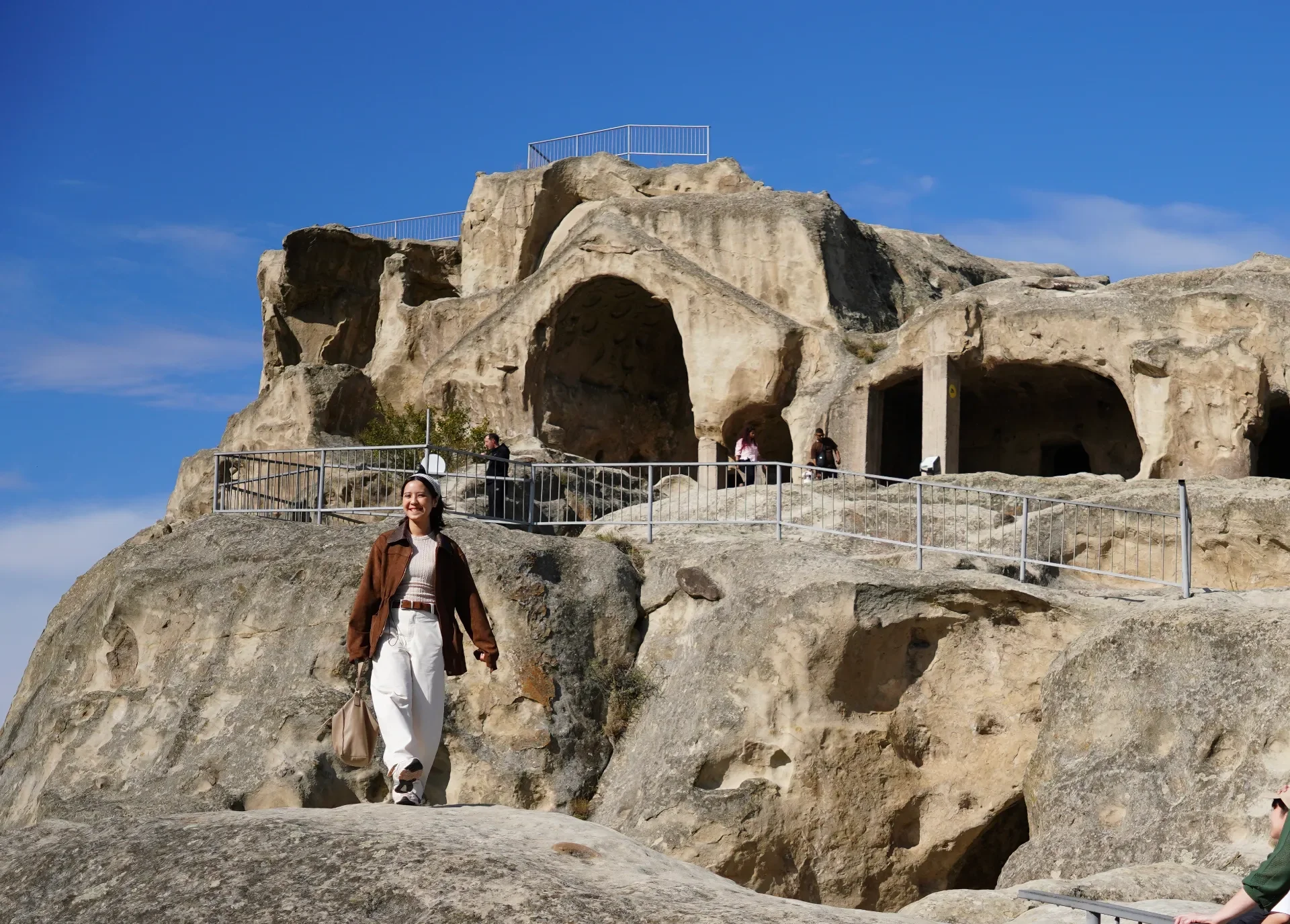Woman smiling in front of ancient cave dwellings carved into large rock formations, with other visitors exploring the site under a blue sky.