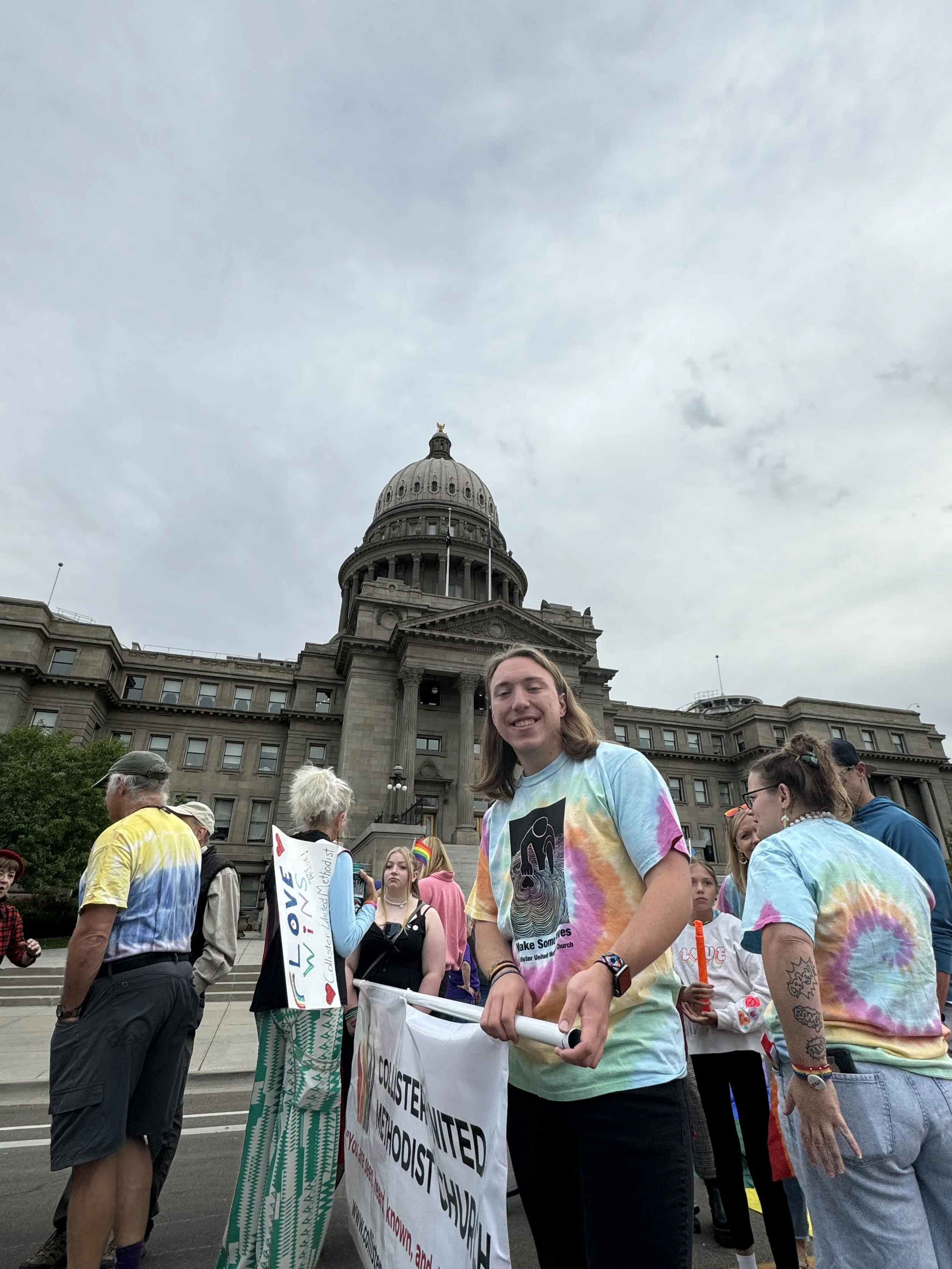 Group of people holding signs in front of a historic government building during a cloudy day.