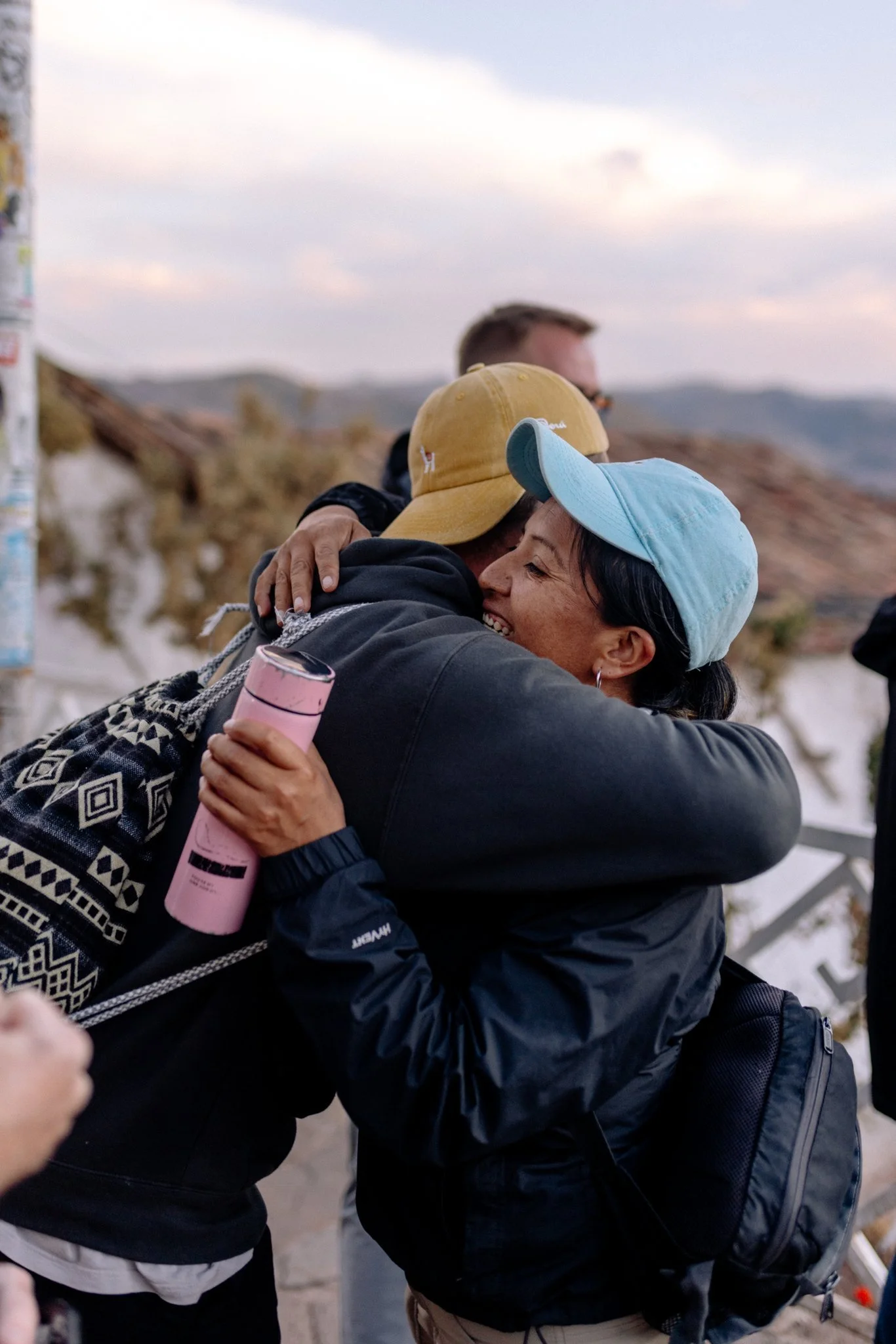 Two women embracing and smiling during a hug outdoors, both wearing caps, one yellow and one light blue, with a background of hills and cloudy sky.