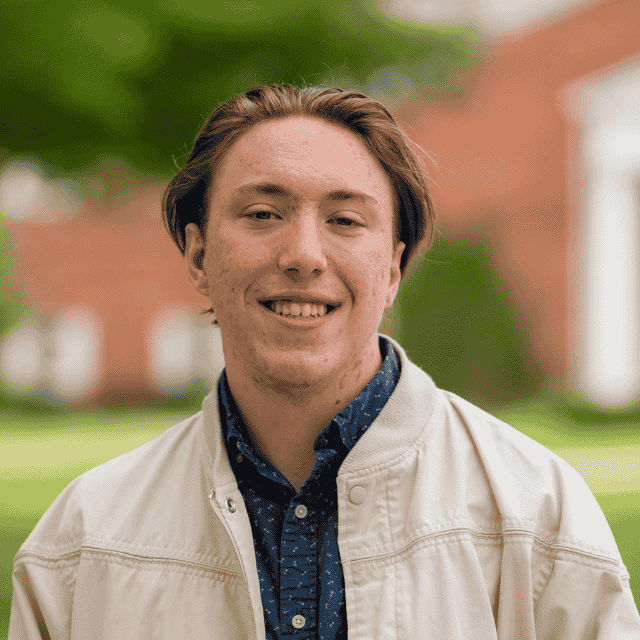 Young man with reddish-brown hair smiling outdoors, wearing a light-colored jacket over a dark, patterned shirt, with a blurred green and brick background.