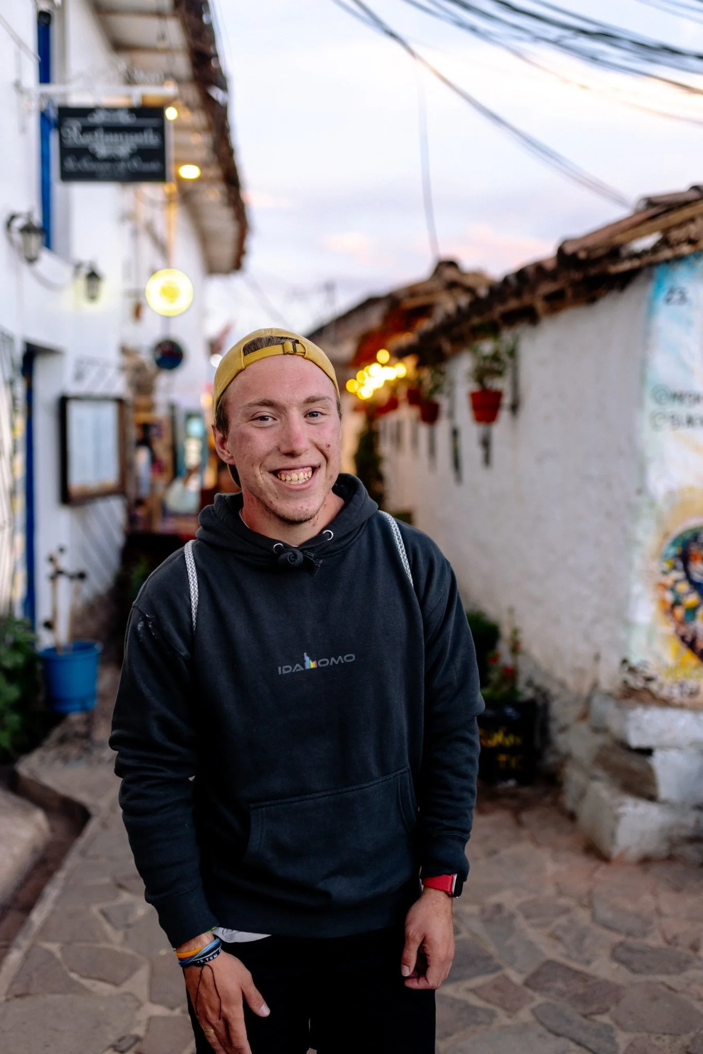 A young man smiling in a narrow street with white buildings, hanging flower pots, and warm glowing lights at sunset.