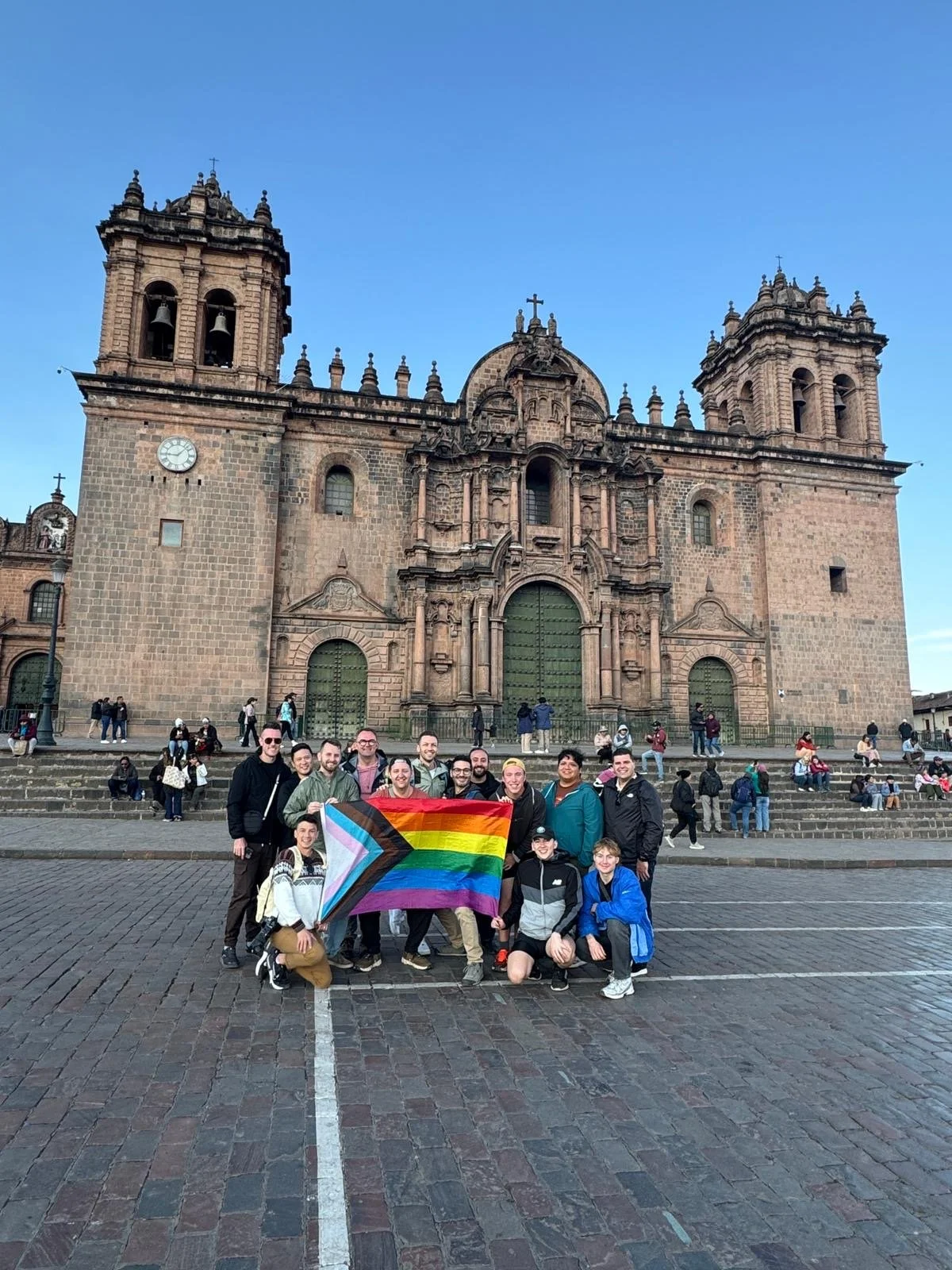 Group of people holding a rainbow flag with a black stripe, pink triangle, and light blue edges in front of a historic stone church with twin bell towers and a large green door, on a cobblestone plaza in the evening.
