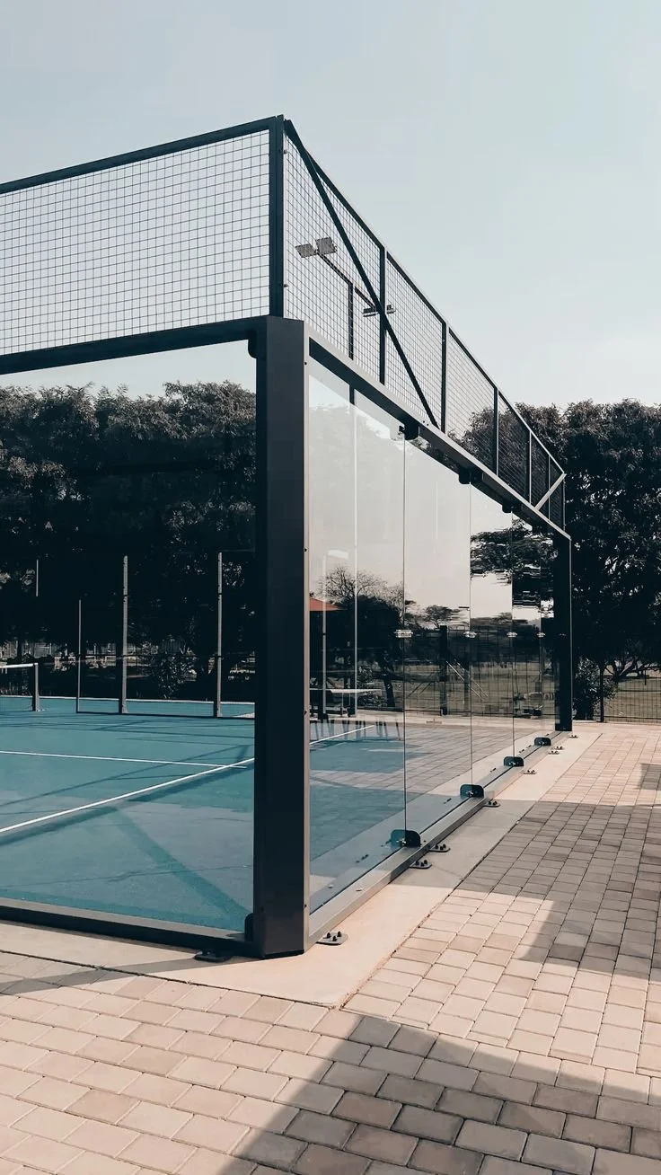 A glass-enclosed paddle tennis court outdoors with metal fencing and a upper-level viewing area in a park or recreational area.