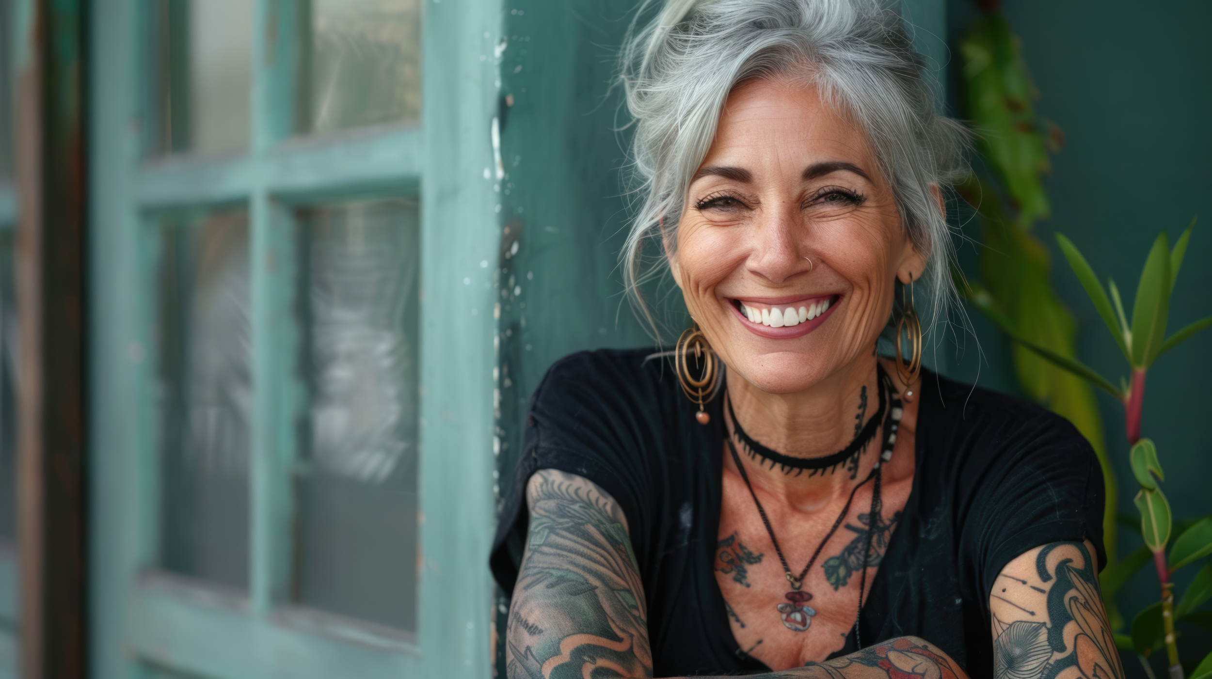 Smiling woman with gray hair, tattoos, and jewelry leaning against a teal door, with greenery in the background.