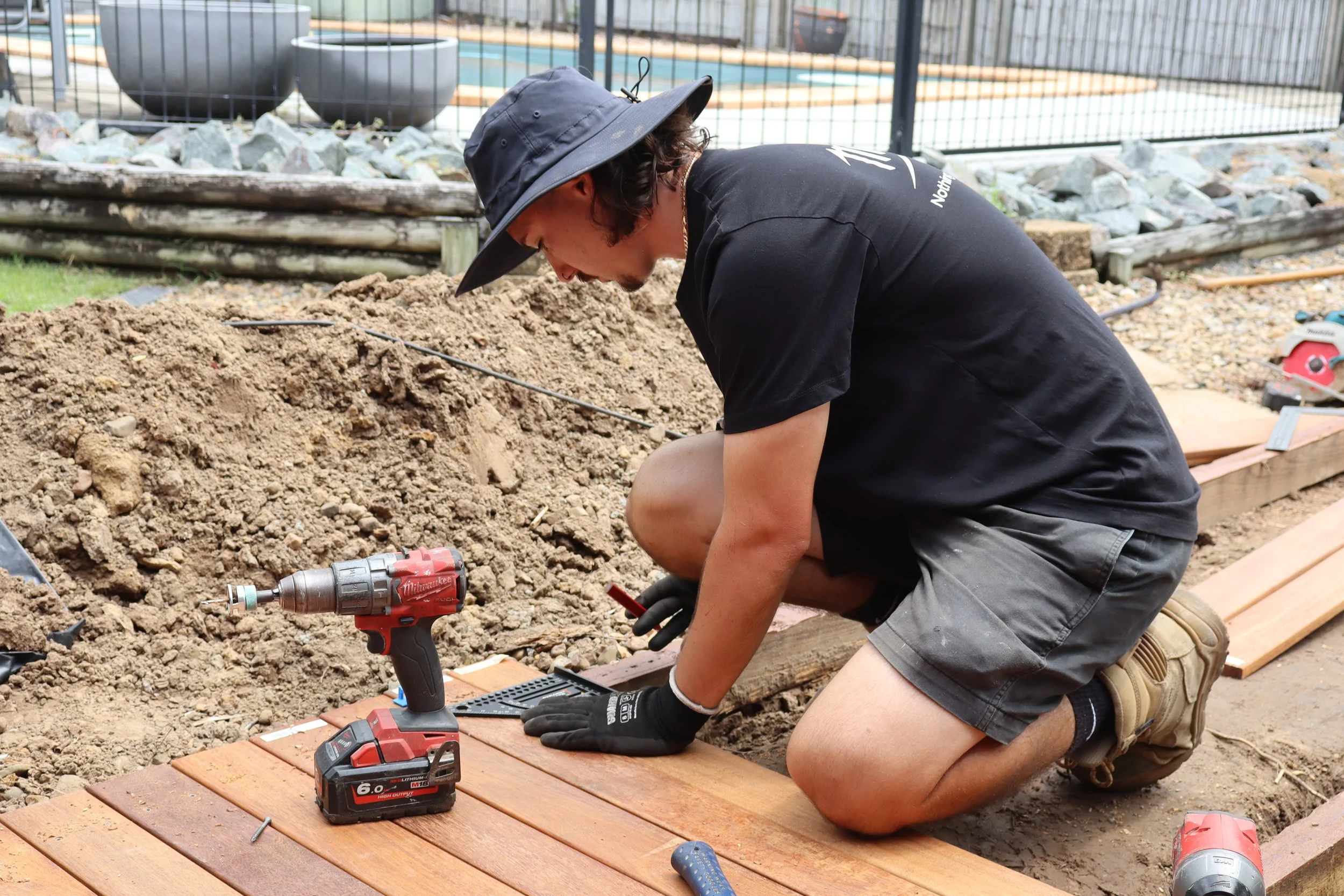 A person kneeling on the ground working on installing a wooden deck, with a cordless drill nearby.