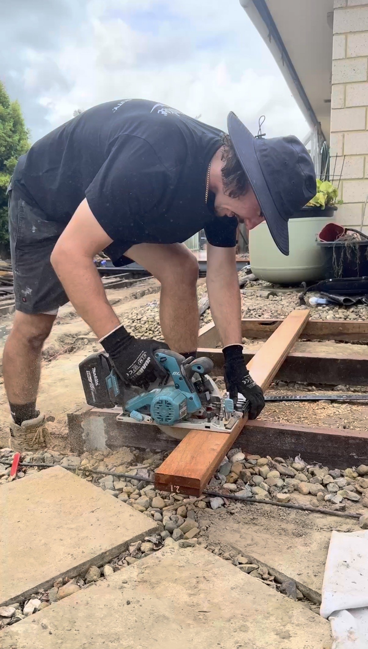 A person using a power saw to cut a wooden board on a construction site outdoors. The person is wearing a wide-brimmed hat, black gloves, and a black t-shirt.