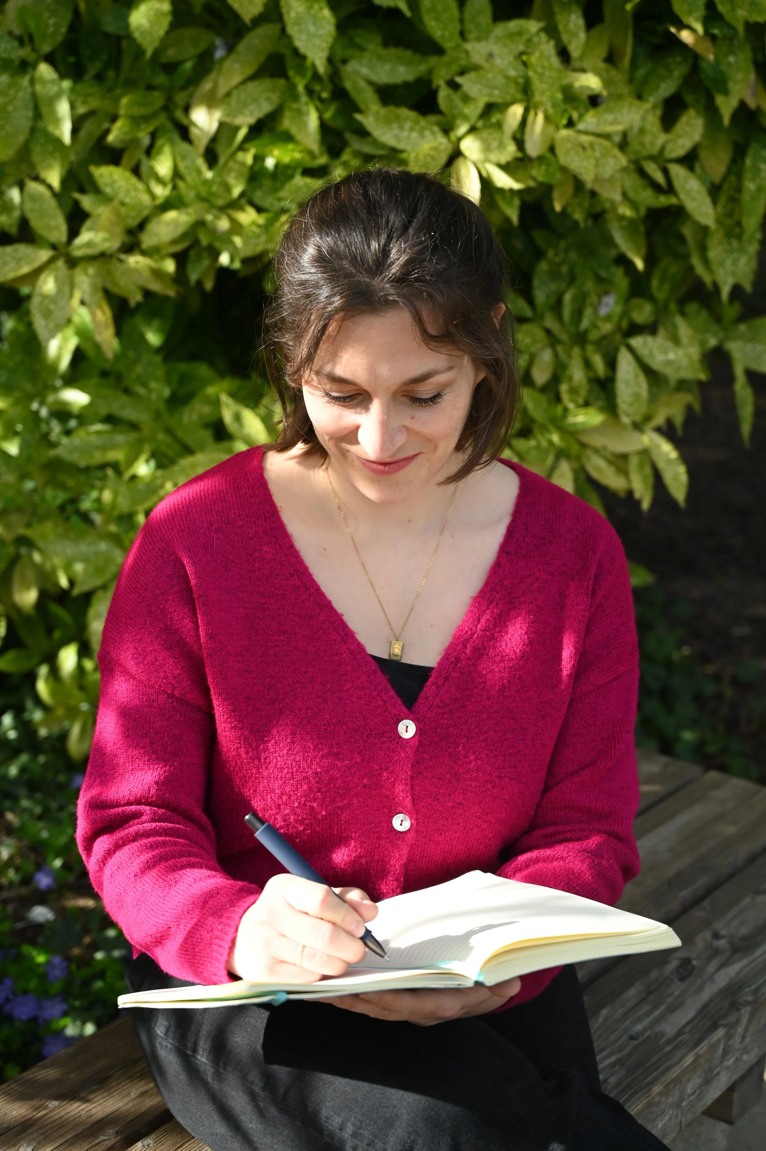A woman in a red cardigan sitting on a wooden bench outdoors, writing in a notebook with a pen, with green foliage in the background.