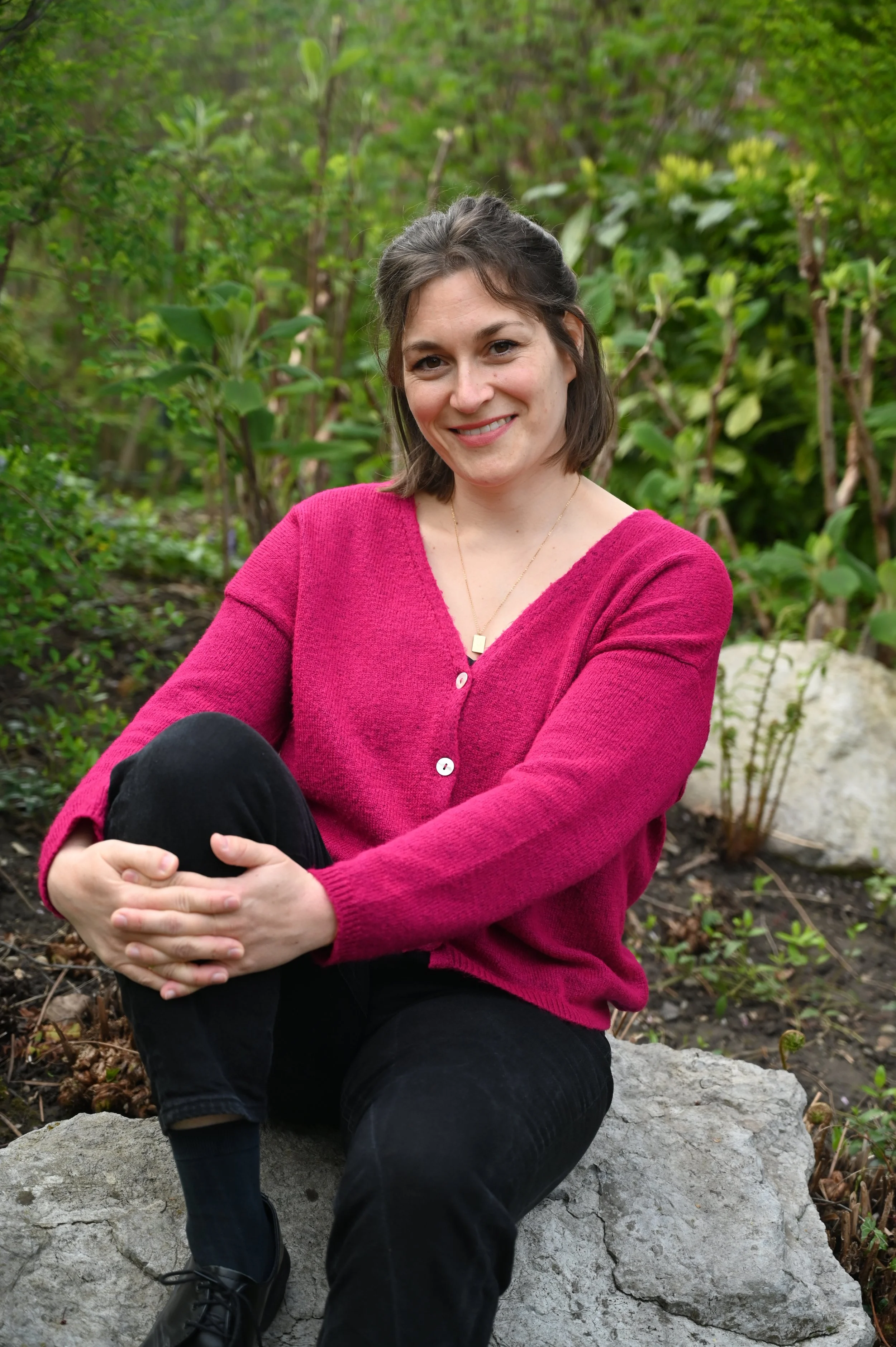 A woman with short brown hair, wearing a pink cardigan and black pants, sitting outdoors on a large gray rock surrounded by green plants and trees, smiling.