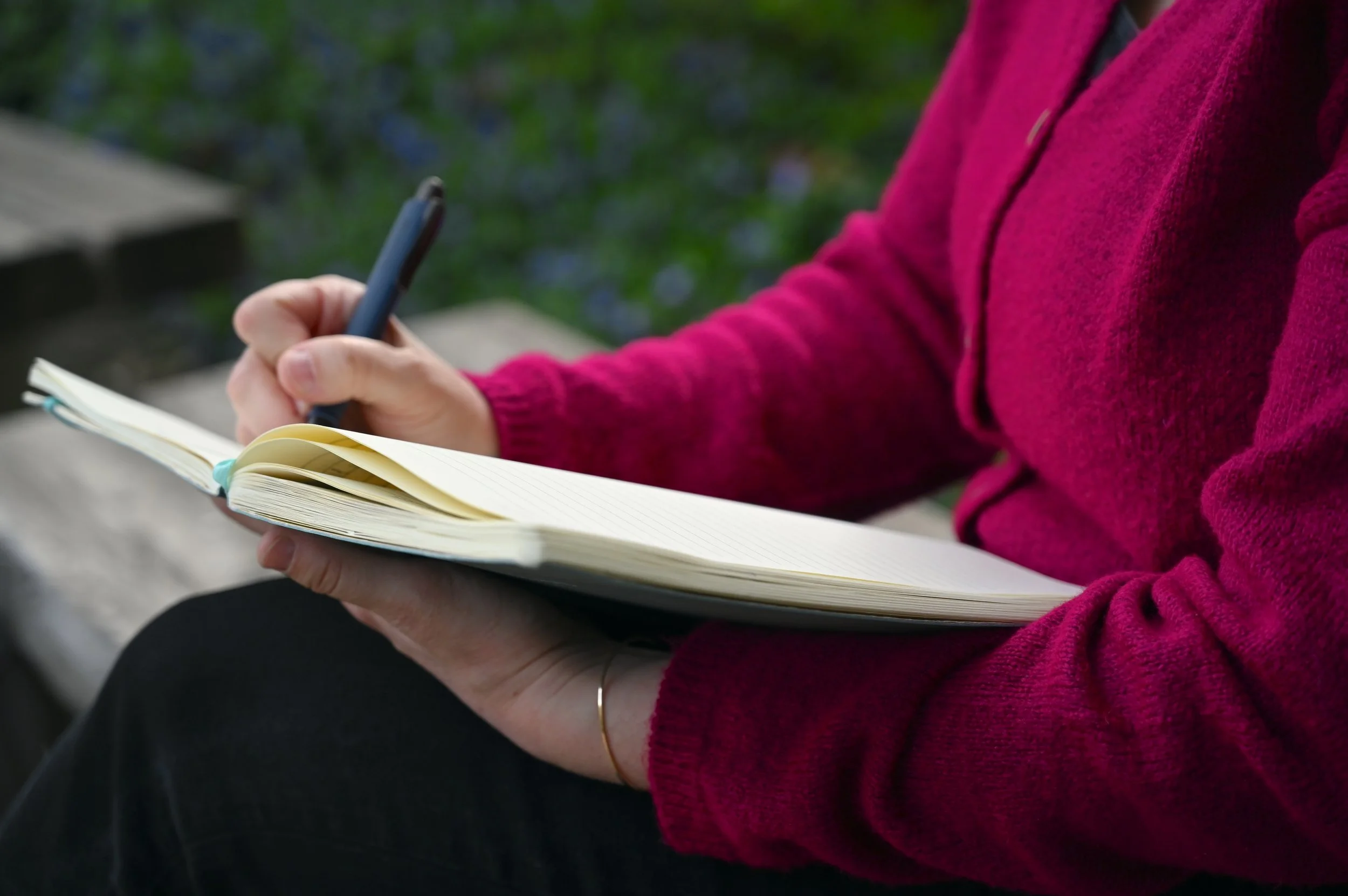 Person wearing a pink sweater, sitting outdoors on a wooden bench, writing in a cream-colored notebook with lined pages.