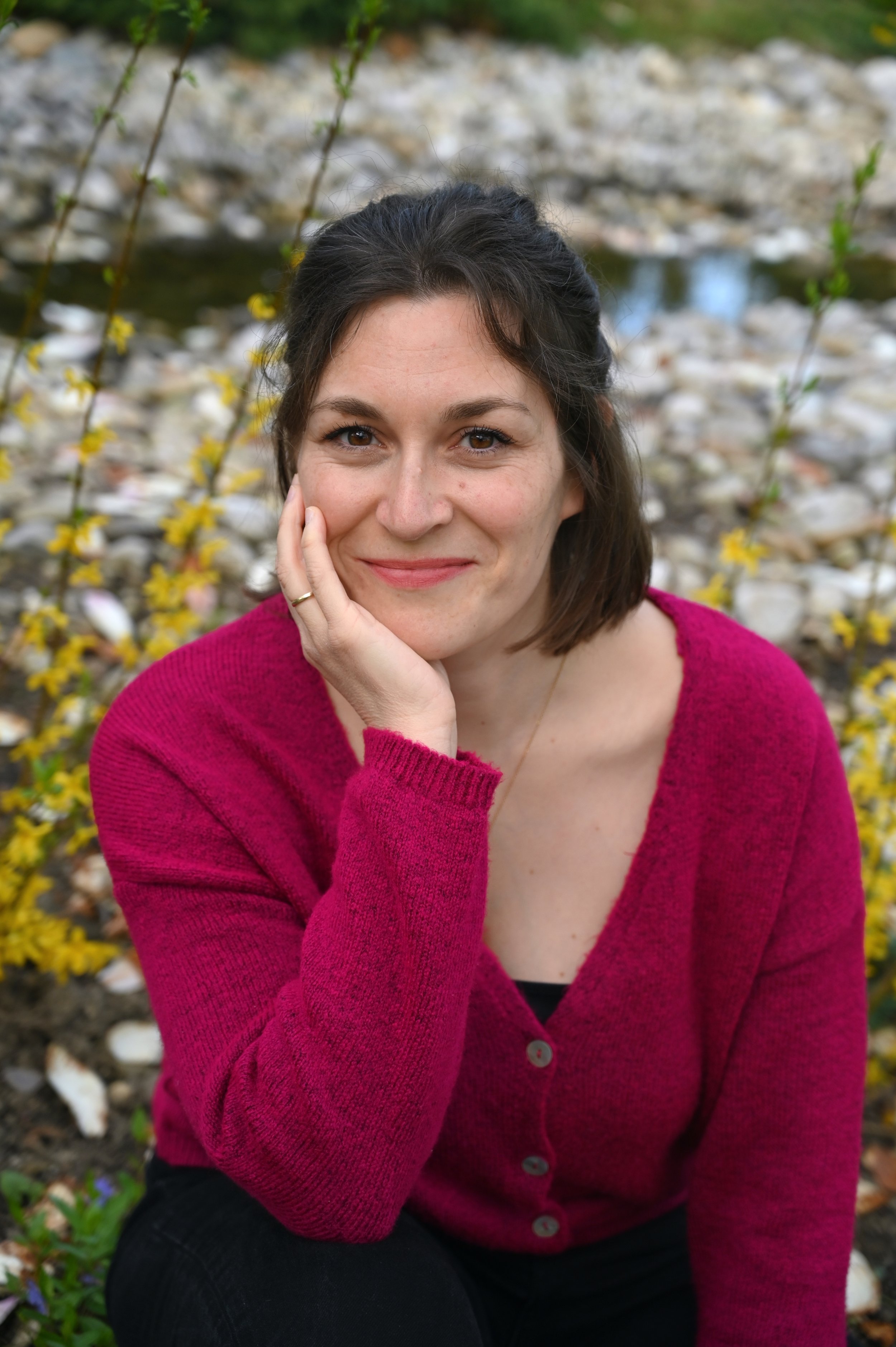 A woman with shoulder-length brown hair, smiling, wearing a pink sweater, sitting outdoors near yellow flowering bushes with a rocky stream in the background.