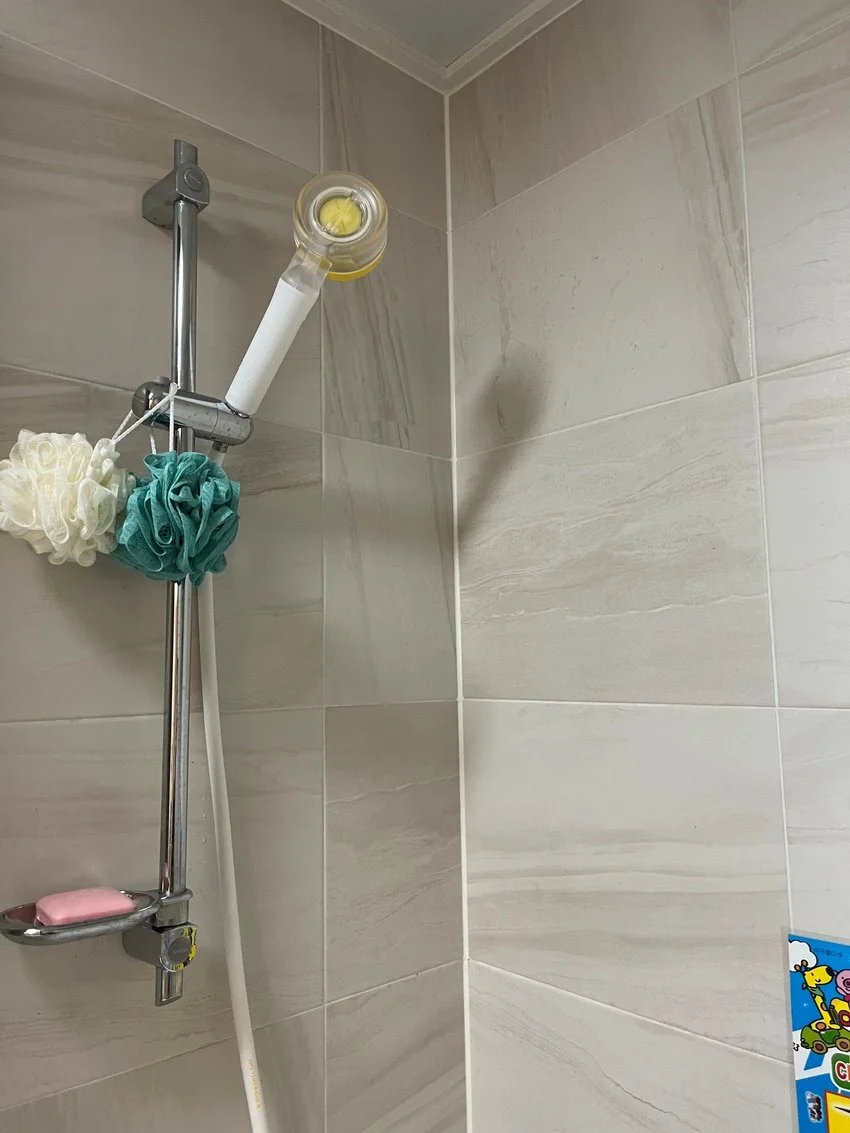 A shower corner with a metal shower rail, a white shower hose, and multiple sponge bath poufs hanging on the rail, including a beige, teal, and a small pink soap bar on a soap dish.