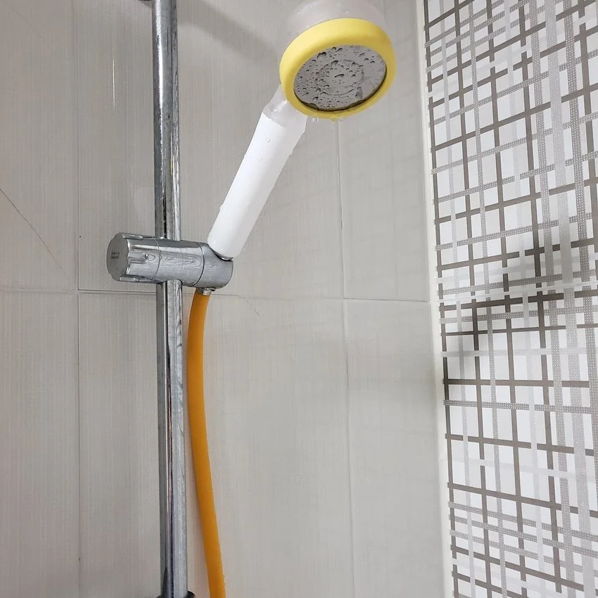 A showerhead attached to a wall with a yellow rim, connected to a flexible orange hose, in a bathroom with tiled walls.