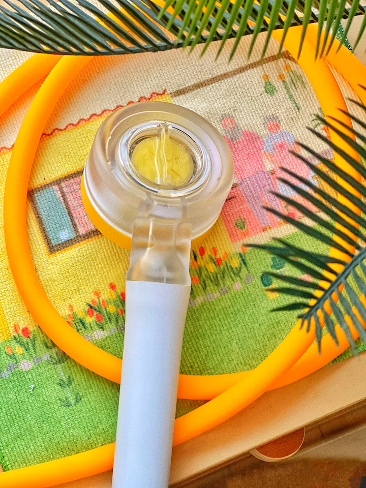 A UV flashlight with a white handle, a yellow and gray body, and a circular transparent top, placed on a colorful cross-stitch cloth with a house and flowers, and surrounded by green palm leaves and an orange plastic ring.