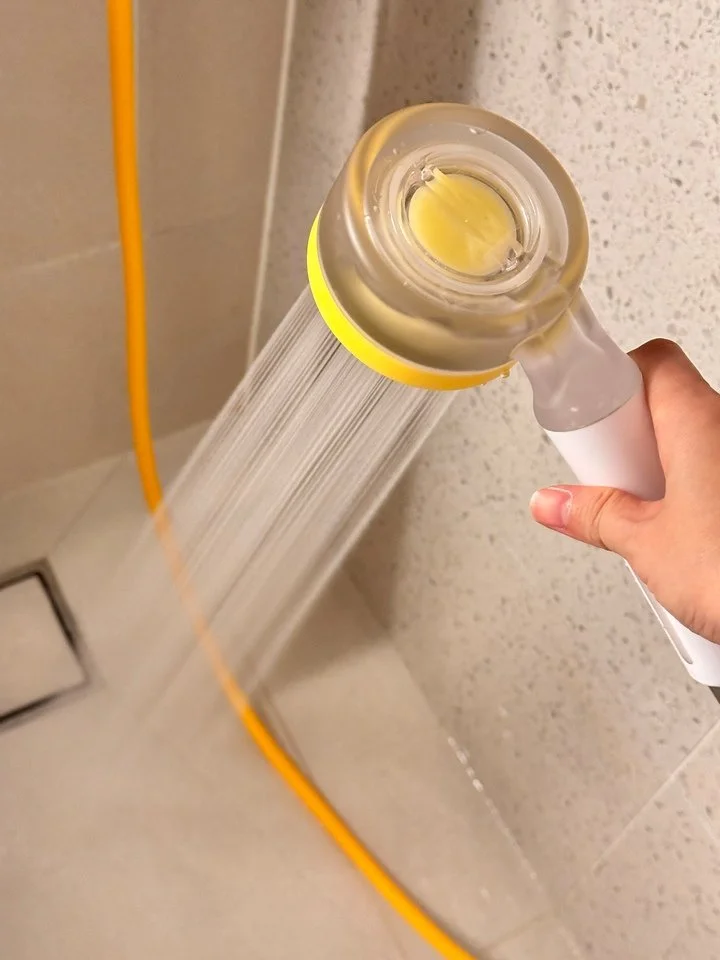 A person is holding a showerhead with a stream of water coming out against a tiled bathroom wall.