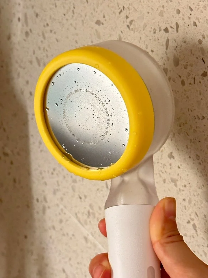 A hand holding a yellow and white portable fan with a circular metal grill, with water droplets visible on the grill and hand. The surface underneath is a speckled countertop.