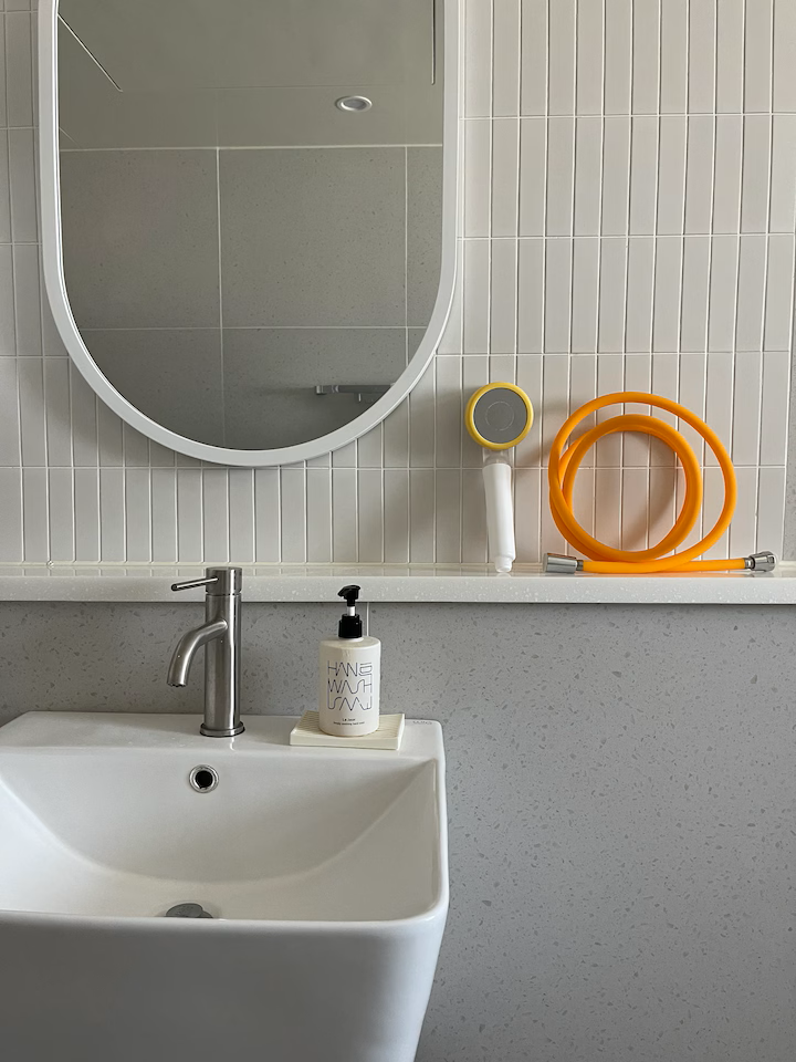 Bathroom counter with a white sink, a soap dispenser, a mirror, and cleaning tools including a spray nozzle, orange hoses, and a gray and yellow scrubber.
