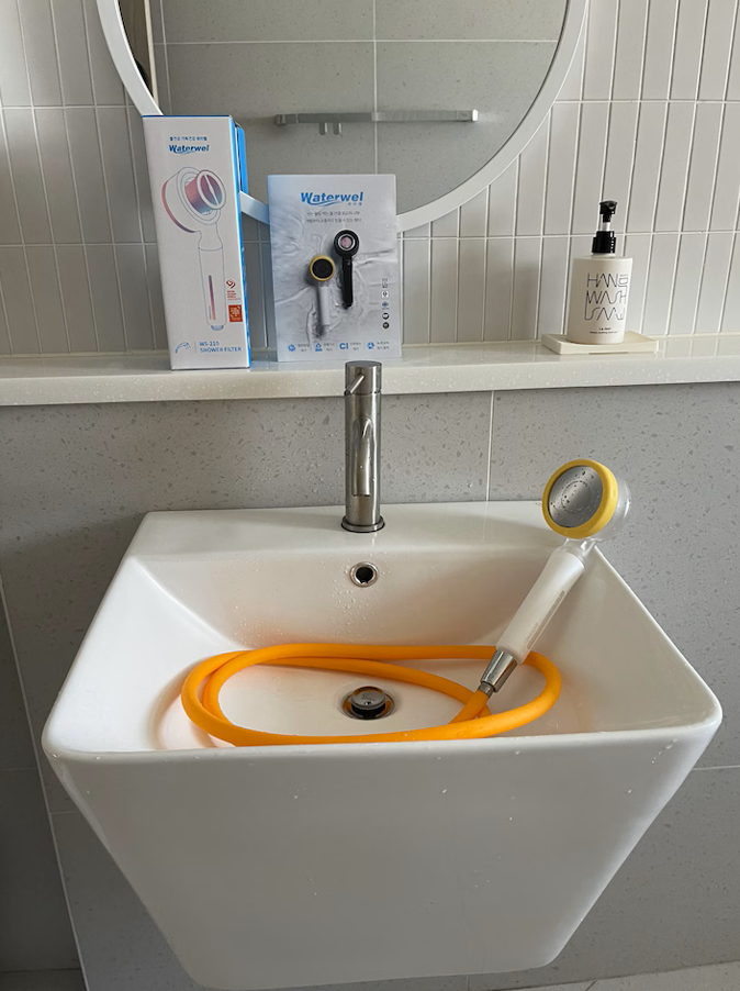 A white bathroom sink with an orange shower hose and handheld shower head inside it. There is a mirror above the sink, a bottle of hand soap, and some visitor door handles on the wall.