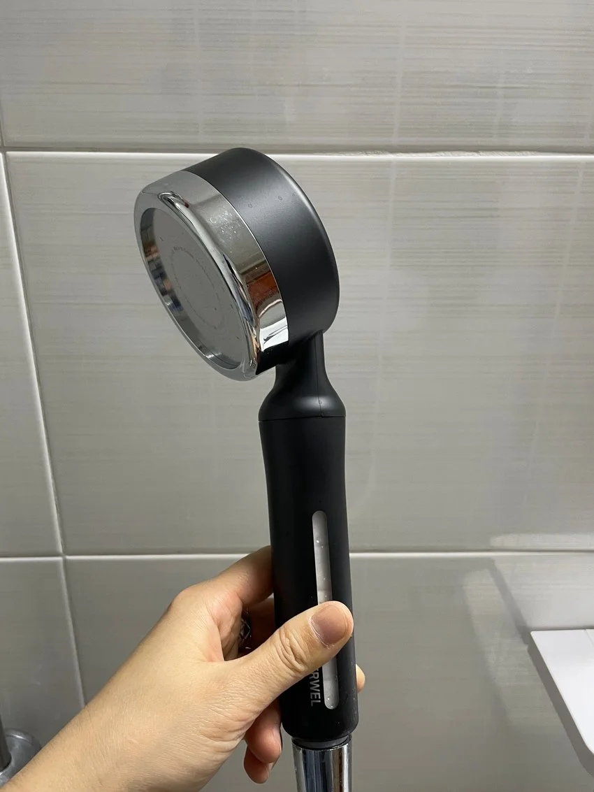 A person holding a black and silver handheld shower head against a tiled bathroom wall.