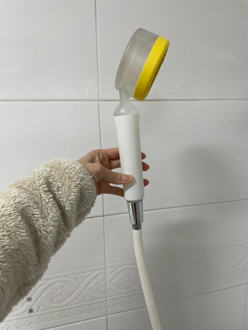 A person's hand holding a handheld bidet sprayer with a white handle, silver connection, yellow and transparent spray head, against a white tiled wall.